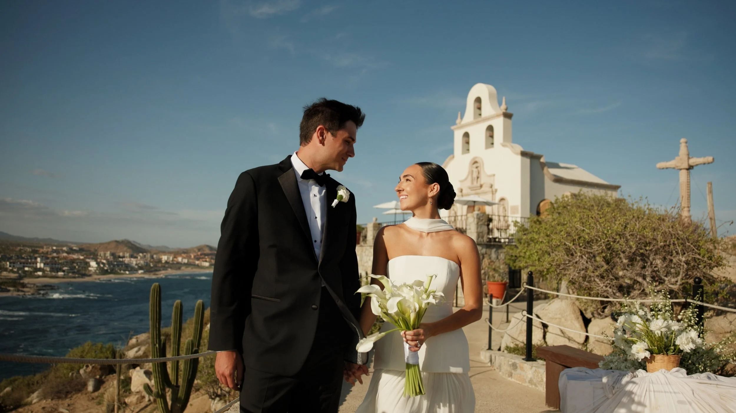 Bride and groom embracing outdoors, groom in tuxedo and bride in lace wedding dress with veil, palm trees in the background.