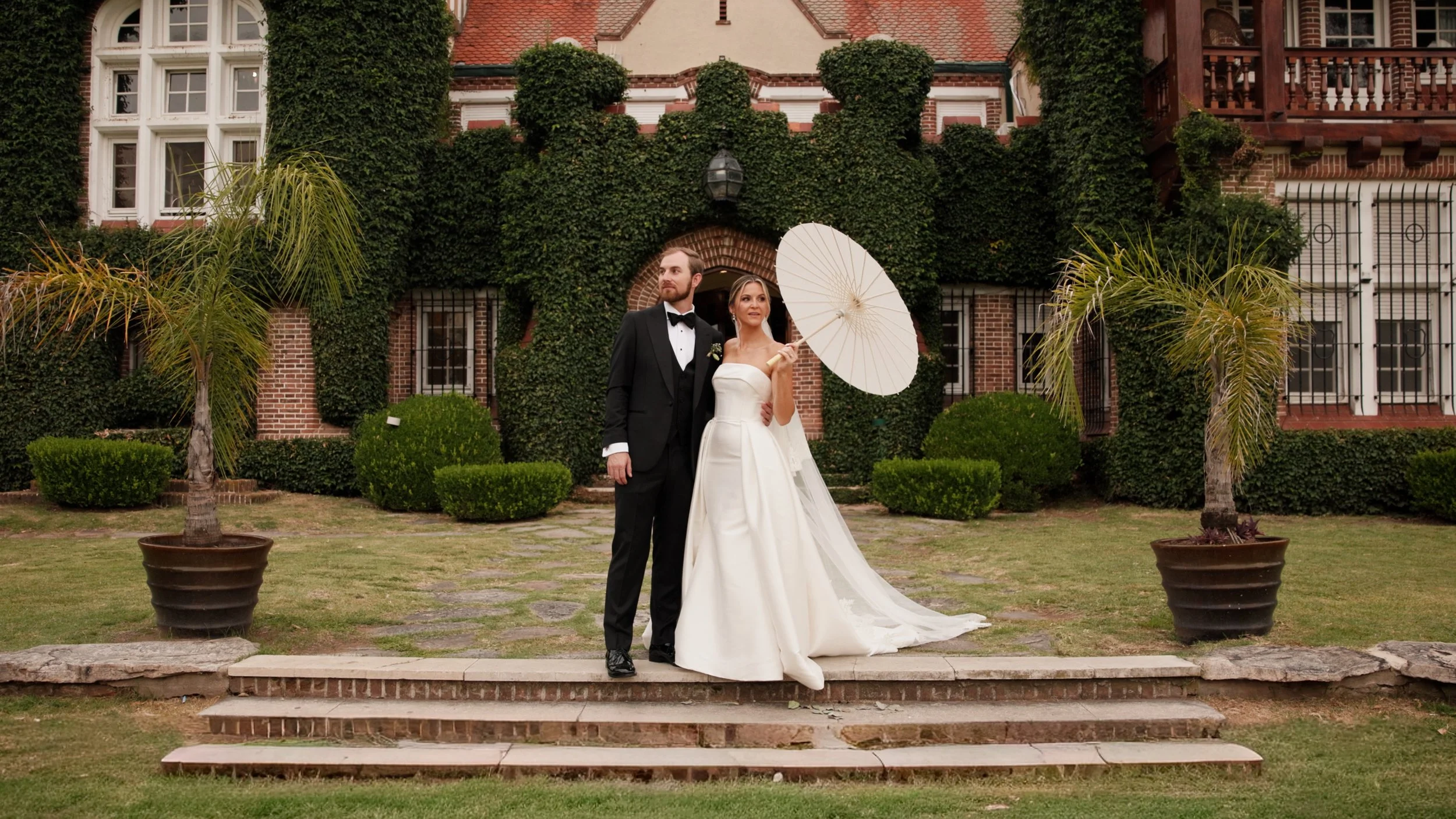 Bride and groom in formal attire embracing outdoors at sunset, with trees and a field in the background.