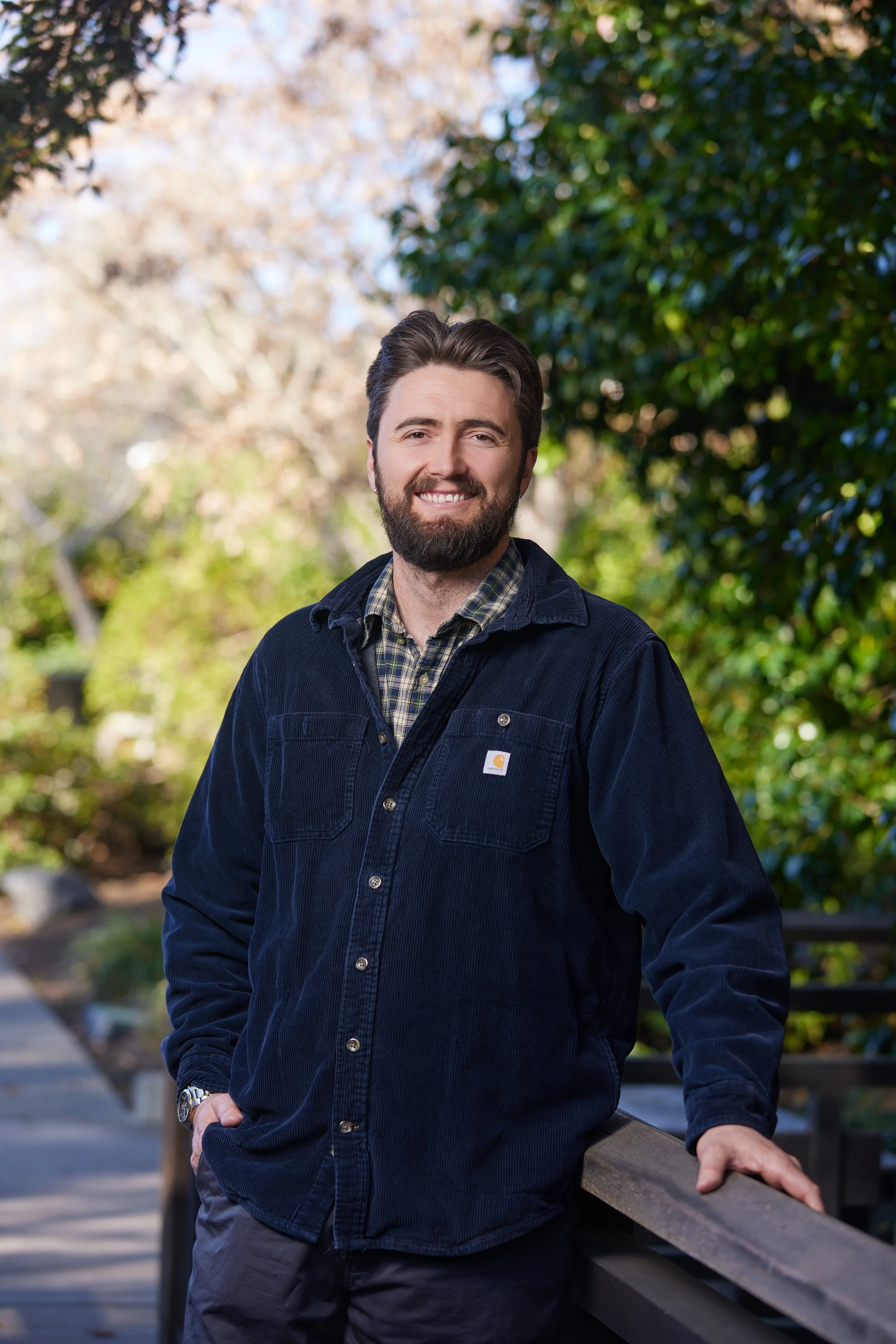 A man with dark hair and a beard smiling outdoors, wearing a dark blue jacket over a plaid shirt, standing by a wooden railing on a pathway surrounded by trees.
