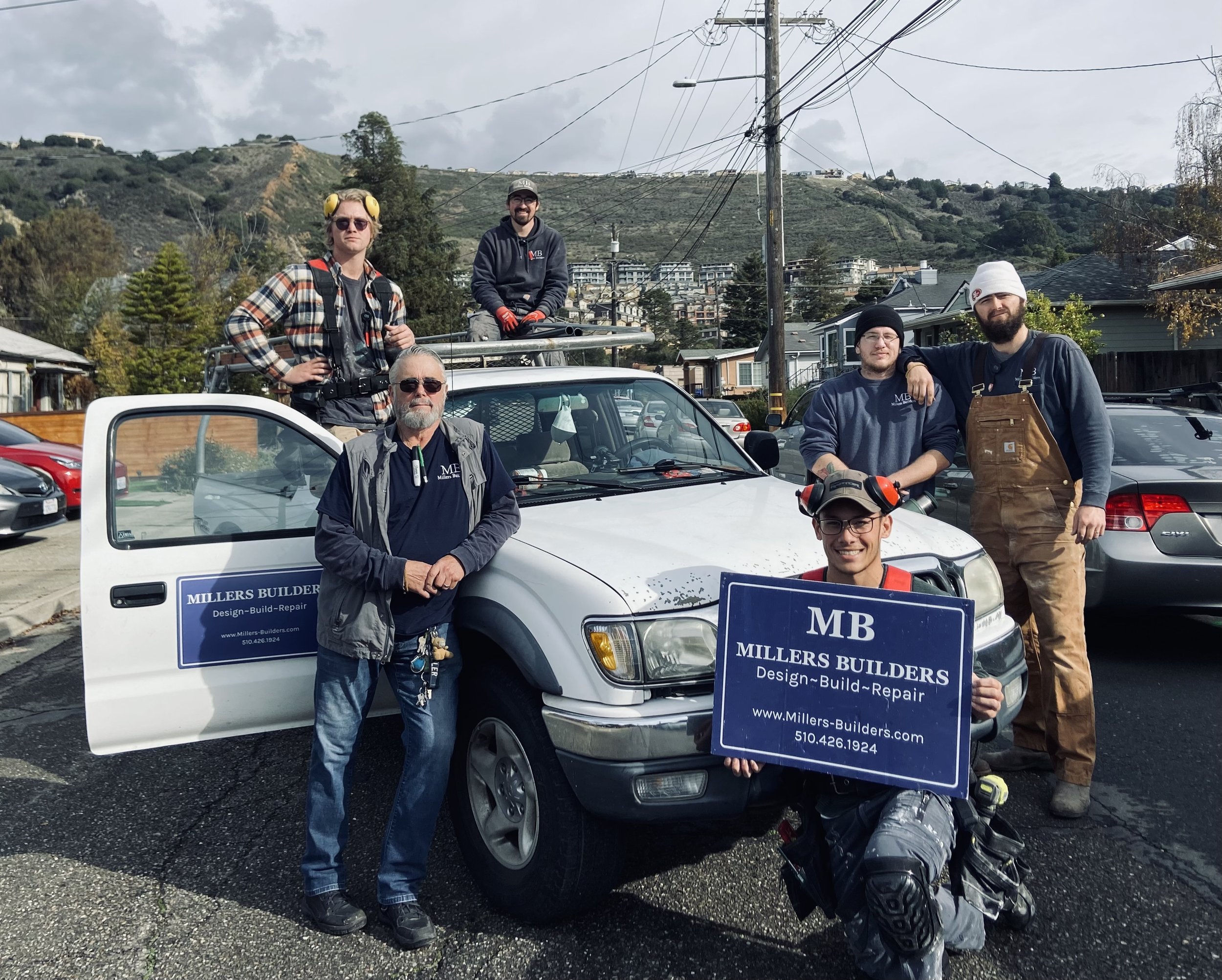Group of five men standing and sitting in front of a white work truck with blue signs, in a parking lot with rocky hills and residential buildings in the background, overcast sky.