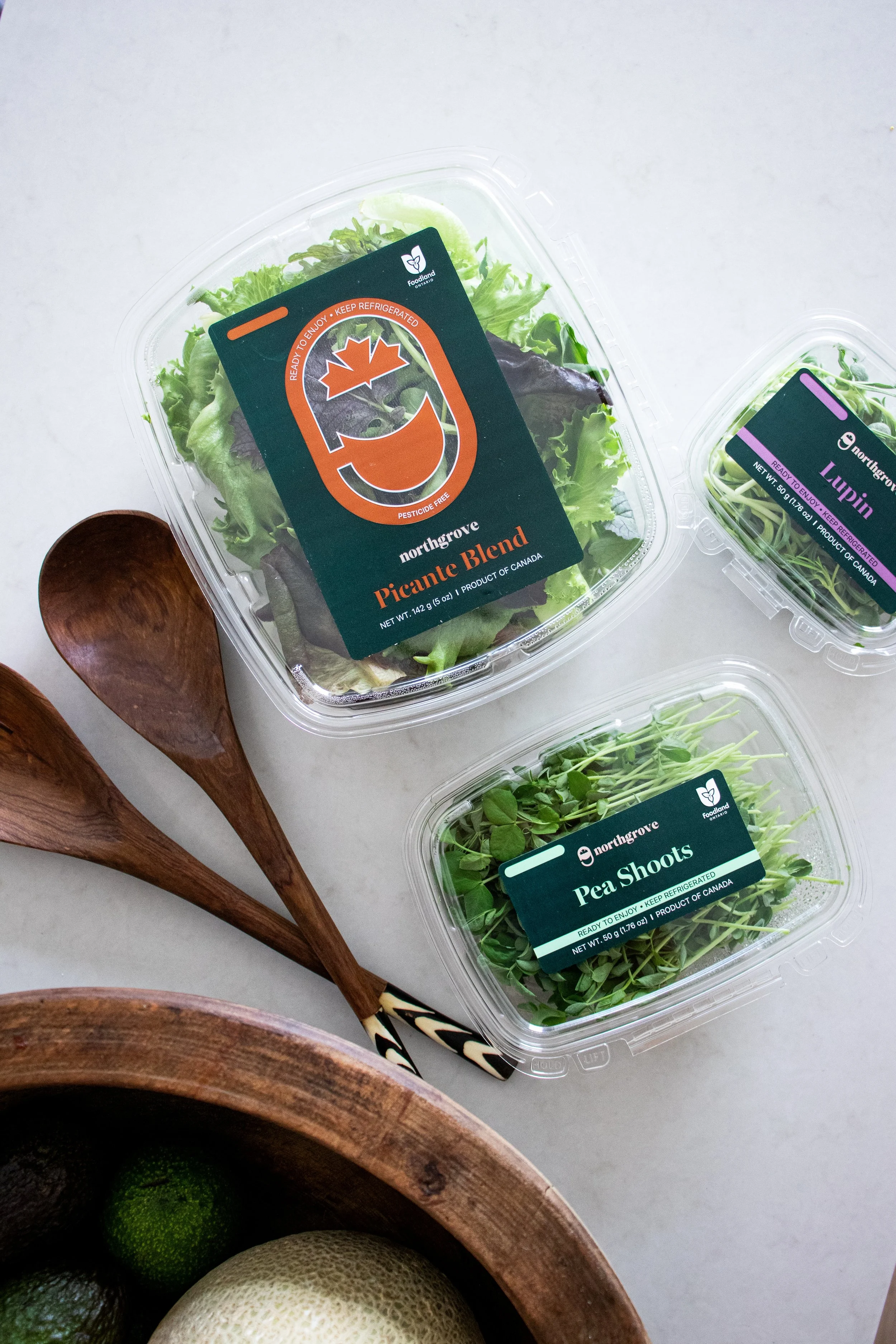 Assorted fresh vegetables in plastic containers, including green pea shoots, mixed greens, and microgreens, with wooden bowls containing melons and limes nearby.