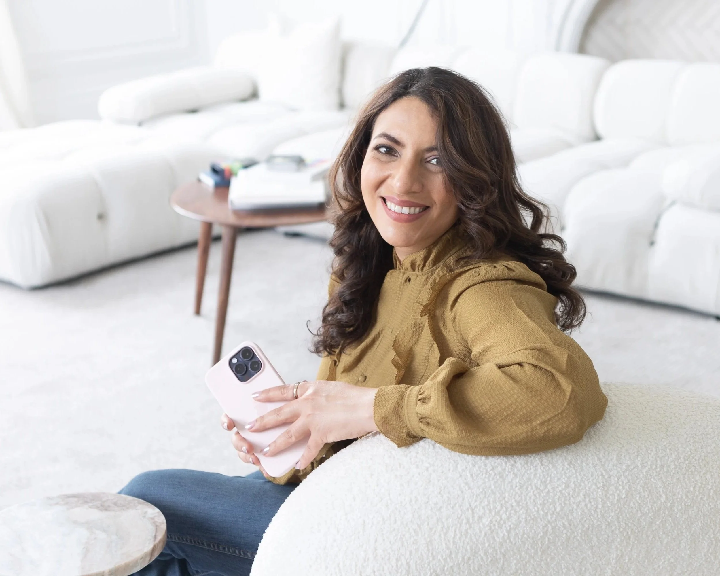 A woman with dark, curly hair smiling and holding a pink smartphone while sitting on a white sofa in a bright living room.