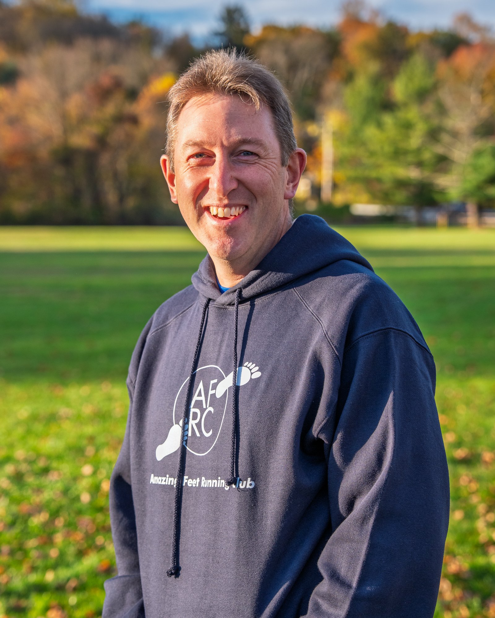 A man smiling outdoors in a park during fall, wearing a navy hoodie with a logo and text that reads 'Amazing Feet Running Club', with trees and grass in the background.