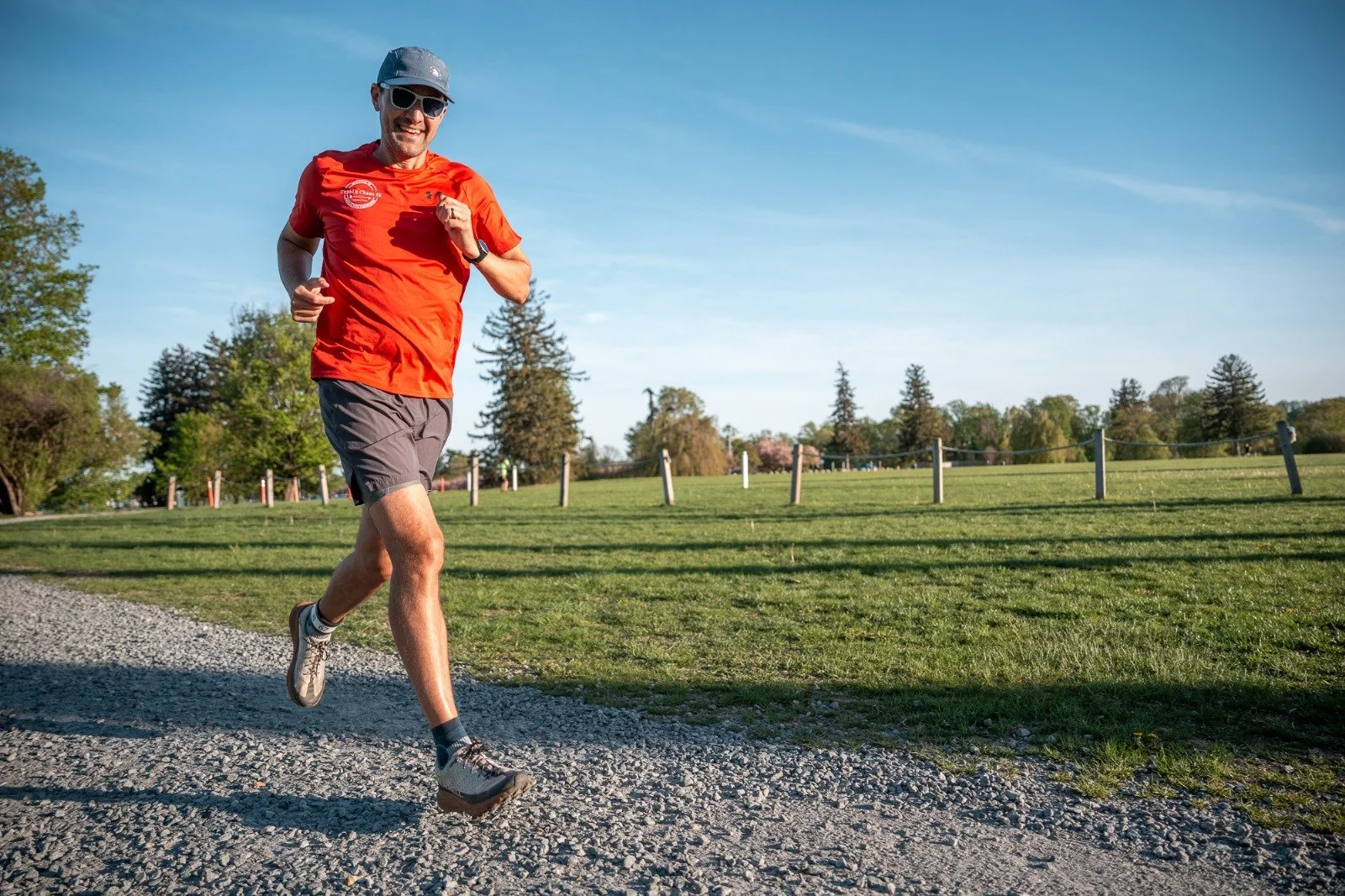 A man running outdoors during a marathon, wearing a red shirt, gray shorts, sunglasses, a blue headband, and a marathon bib with the name Jeremy, number 31. There is a river and trees in the background, and a logo in the lower left corner for the Mount Desert Island Marathon 2023.