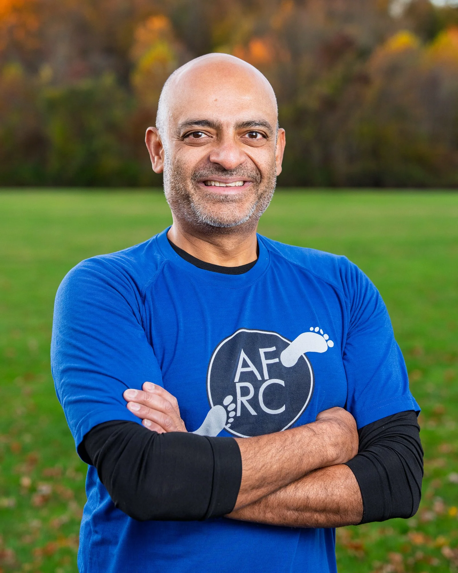 A middle-aged man with a bald head and a short gray beard smiling with arms crossed, wearing a blue T-shirt with a logo featuring a footprint and the letters 'A F R C,' standing outdoors in a grassy field with trees in fall colors in the background.