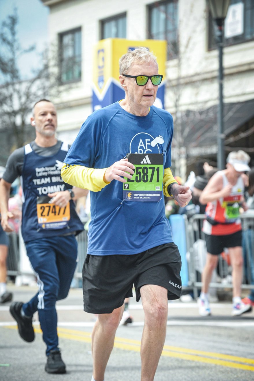 Older man wearing sunglasses, blue shirt, and black shorts running in a marathon, with other runners and city buildings in the background.