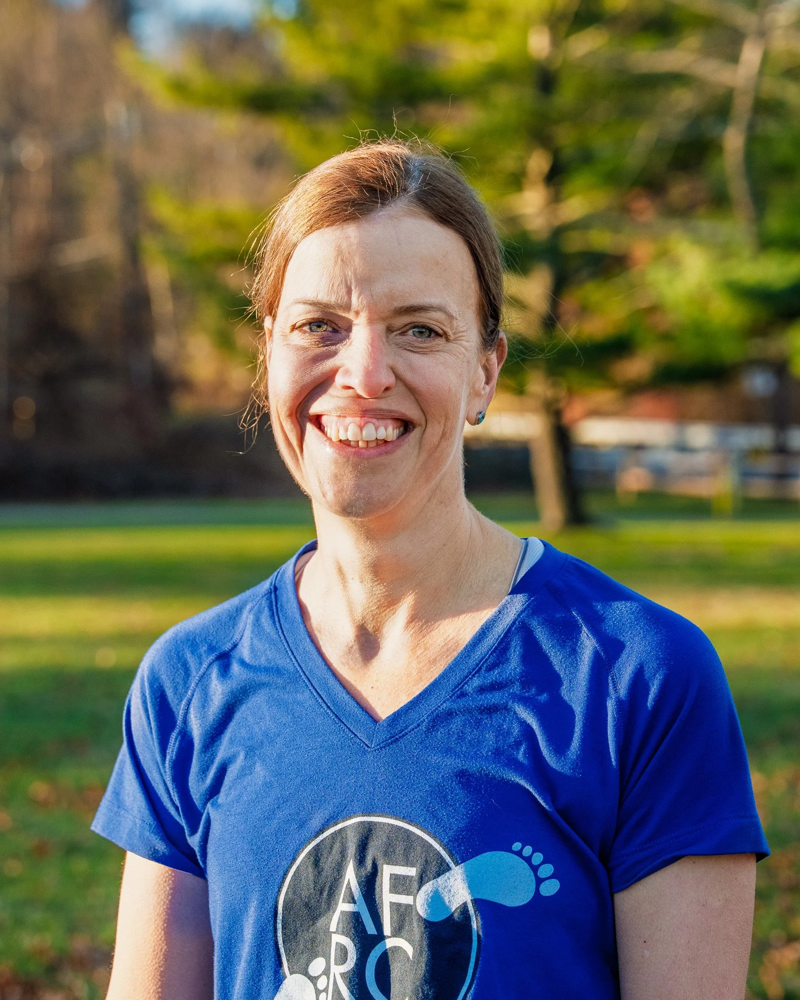 A woman smiling outdoors in a park, wearing a blue athletic shirt with a logo, with trees and grass in the background.