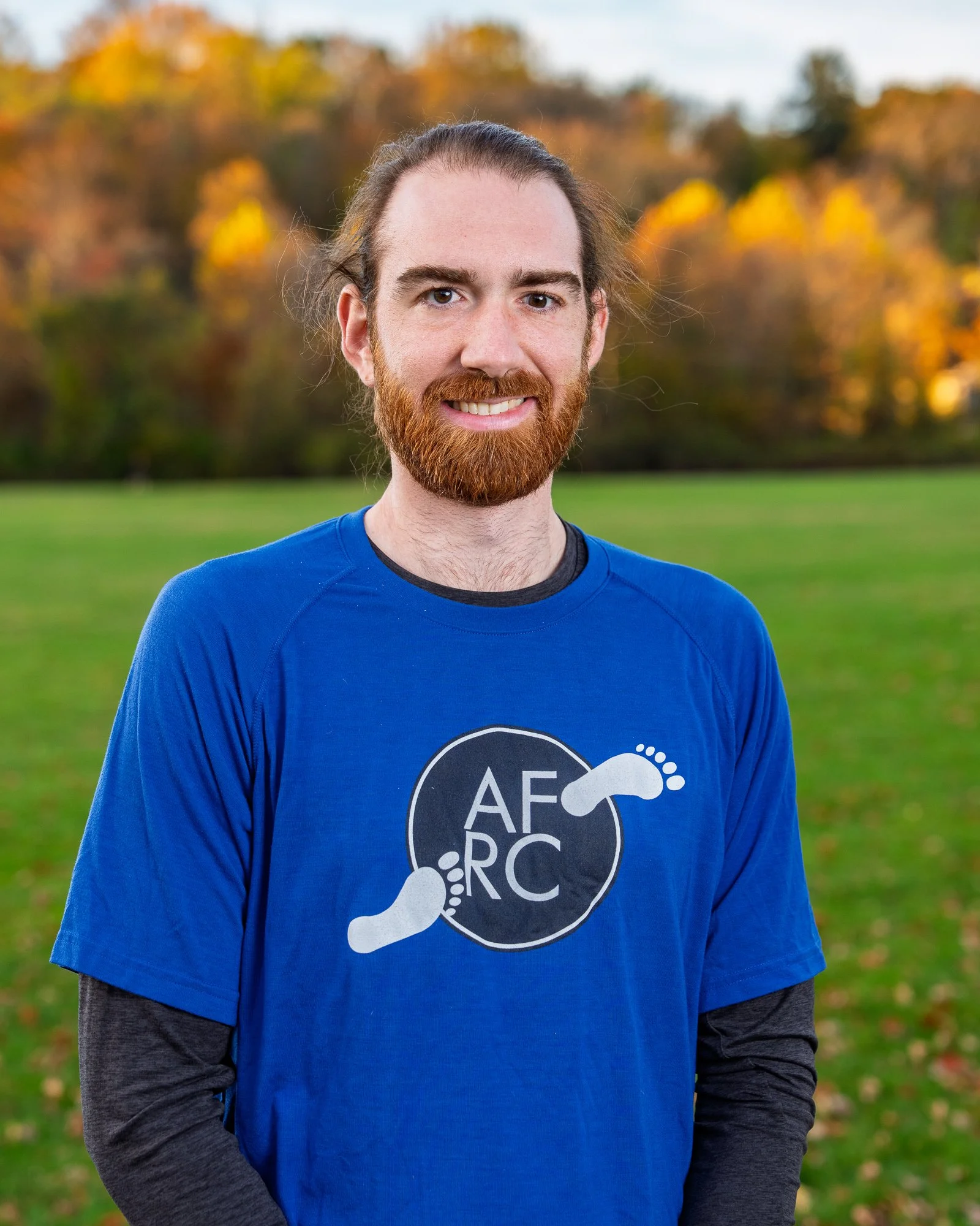 A man with long hair, a beard, and light skin, wearing a blue shirt with a logo, standing outdoors in a grassy area with trees showing fall colors in the background.