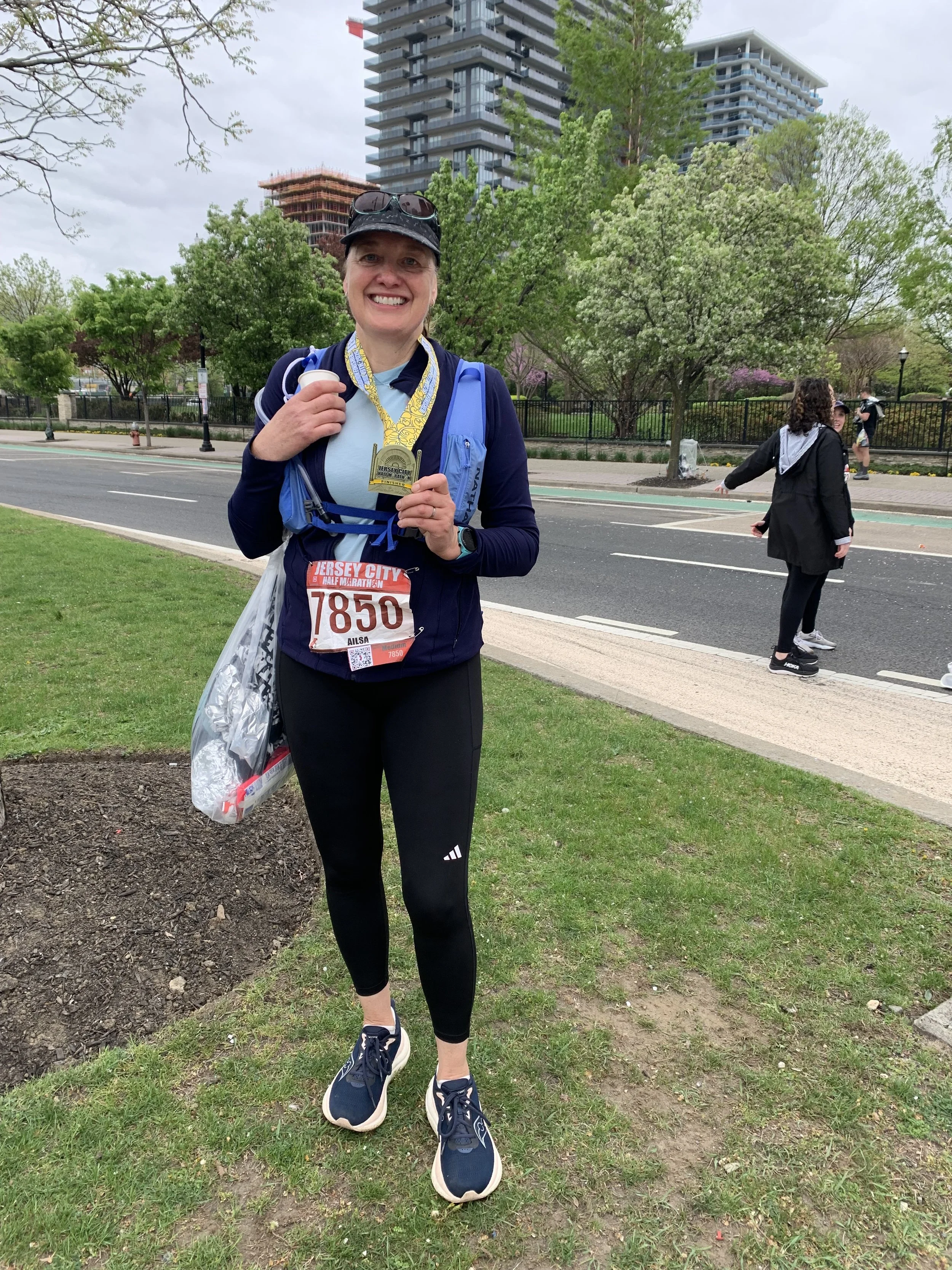 A smiling woman wearing a race bib and medals, holding a water cup, standing on a grassy area after a race, with trees, a sidewalk, and tall buildings in the background.