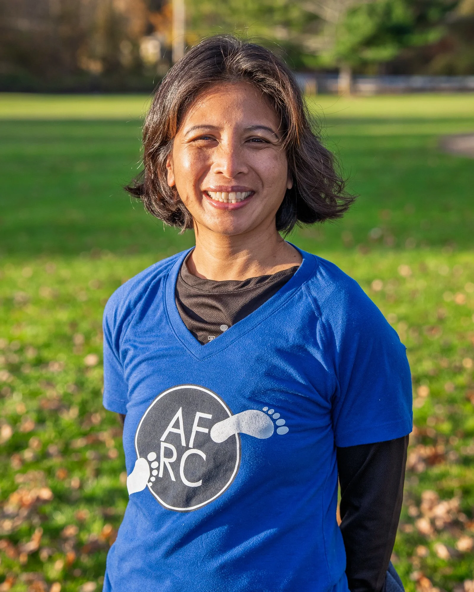 A woman standing outdoors in a park, smiling, wearing a blue T-shirt with the AFERC logo, and black long sleeves underneath, with trees and grass in the background.