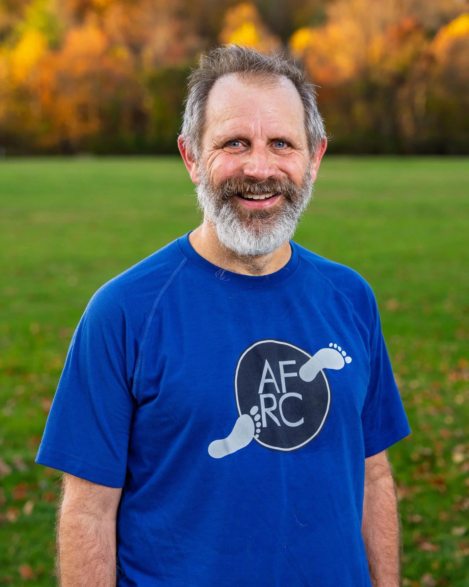 Smiling man with gray beard wearing a blue t-shirt outdoors in a park during fall.