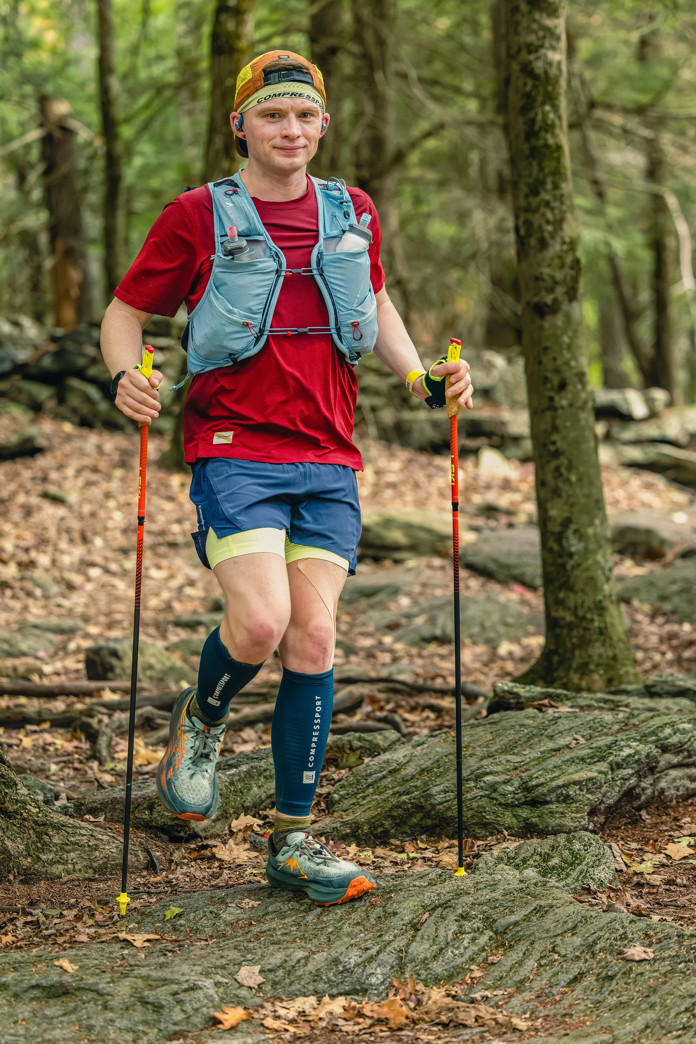 A man trail running on a dirt path through a green forest, wearing a black shirt, orange shorts, a cap, and using trekking poles.