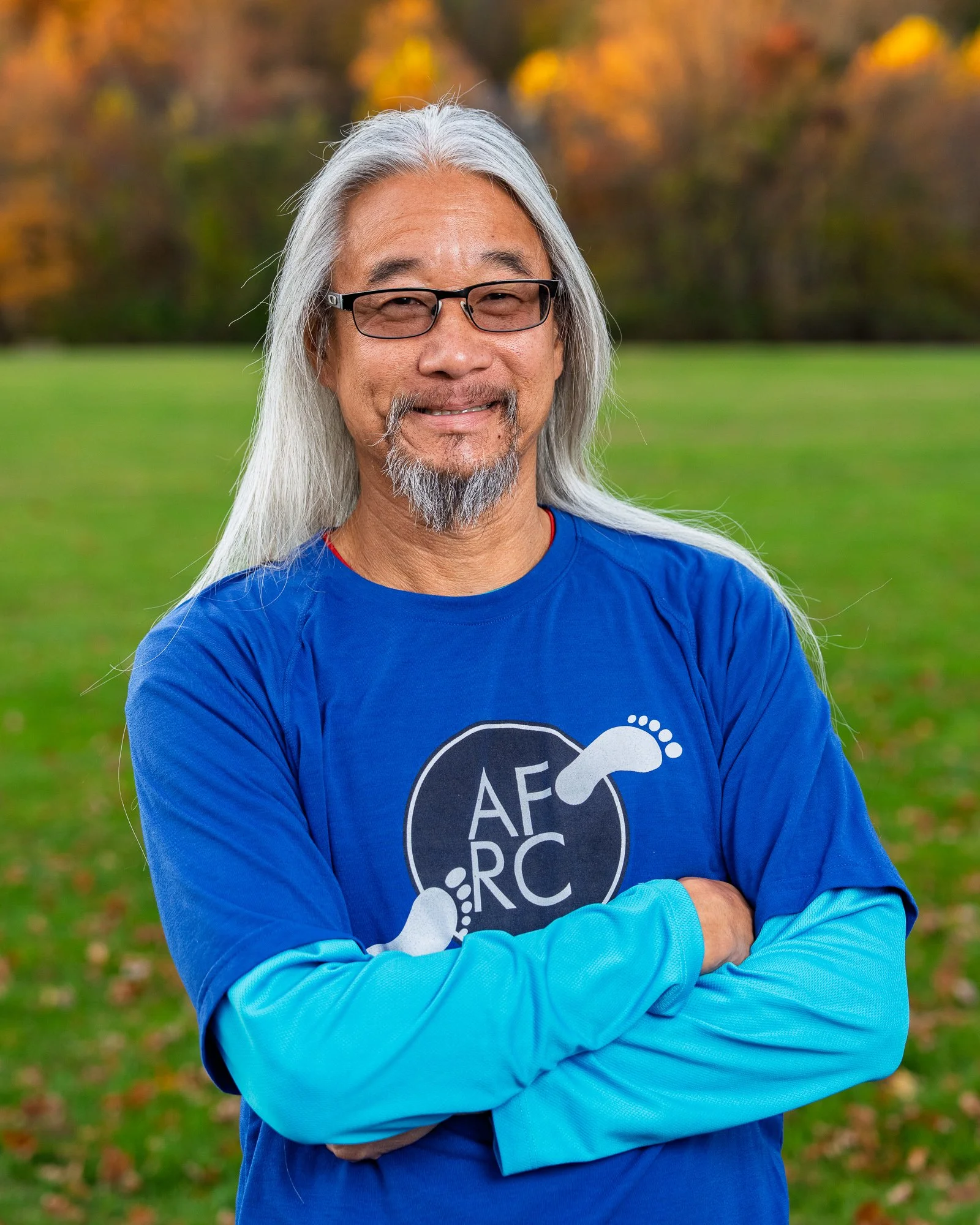 A man with long gray hair, glasses, and a beard standing outdoors with green grass and autumn trees in the background. He is smiling and wearing a blue shirt with a logo featuring a footprint and the text "AF RC".