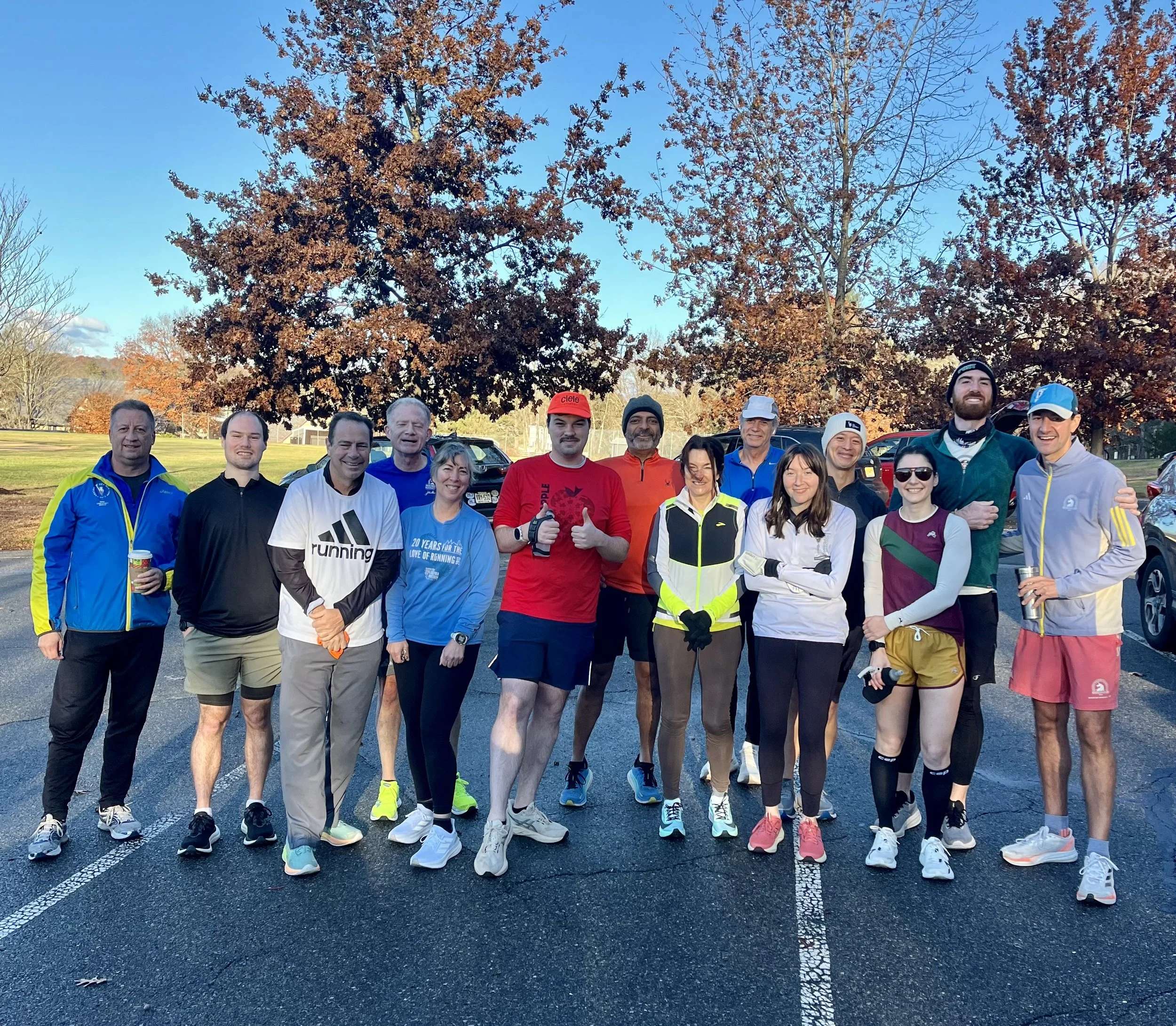 A group of diverse runners standing outdoors in a parking lot during fall, with trees with orange leaves in the background, dressed in athletic gear.