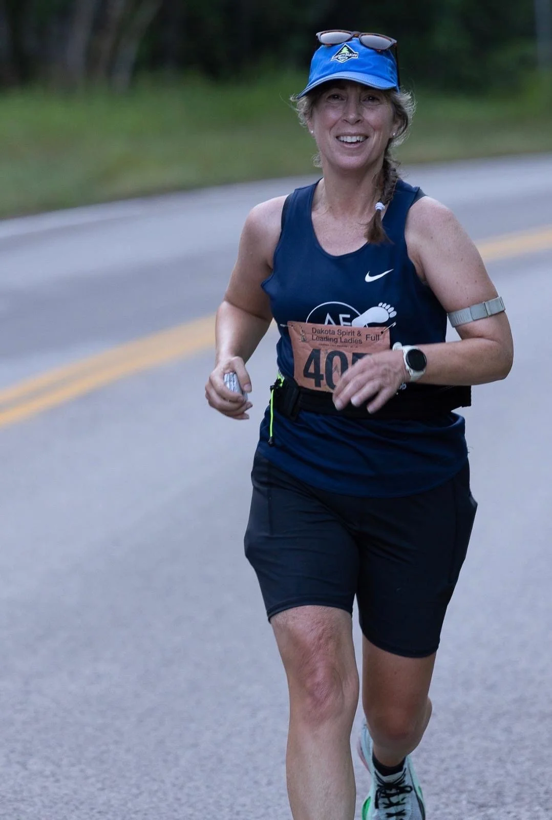 A woman participating in a running race, wearing a purple tank top, black shorts, and yellow running shoes with teal accents, crossing the finish line with a race bib numbered 41 visible on her chest. Several spectators and other runners are in the background.