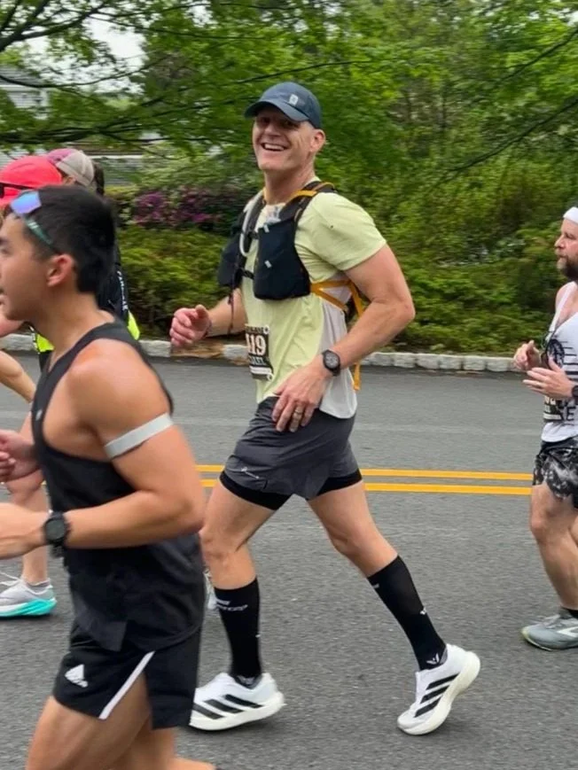 A group of runners participating in a marathon, with a smiling man wearing a gray cap, yellow shirt, black shorts, and running gear, jogging on a road lined with green trees.