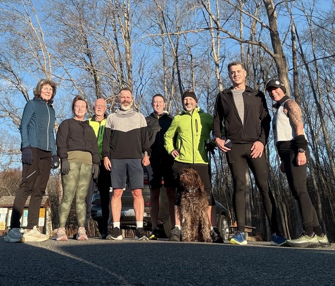 Group of nine people in athletic clothing standing on a paved outdoor area with leafless trees and a blue sky in the background, some with gloves and hats, with a dog sitting in front.