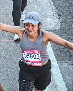 A woman wearing a blue cap and gray athletic pants holds up a medal at the finish line of the Philadelphia Half Marathon, surrounded by fellow runners and event volunteers.