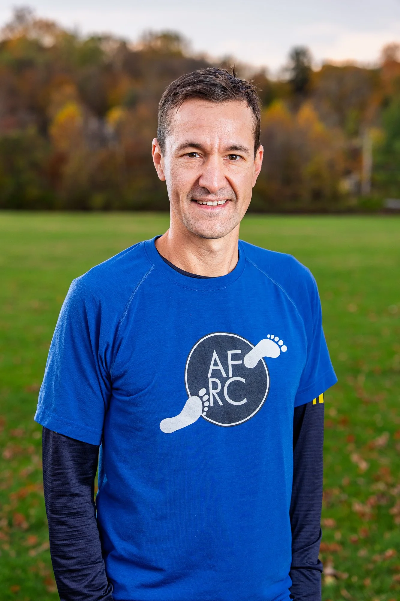 Smiling man wearing a blue athletic shirt with a logo, standing outdoors on a grassy field, with trees in the background.