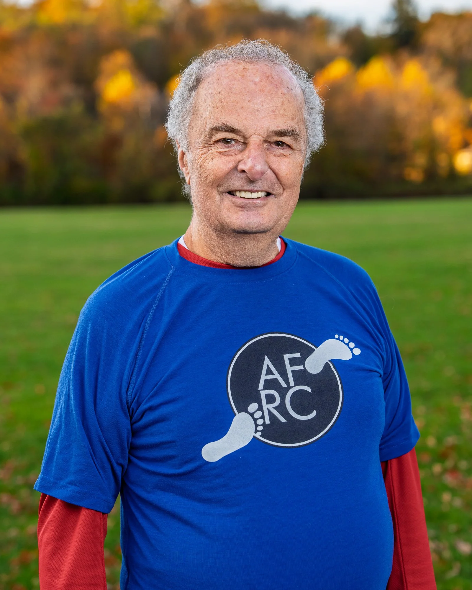Smiling elderly man wearing a blue T-shirt with a logo that includes foot graphics and the text 'AF RC', standing outdoors on a grassy field with autumn trees in the background.