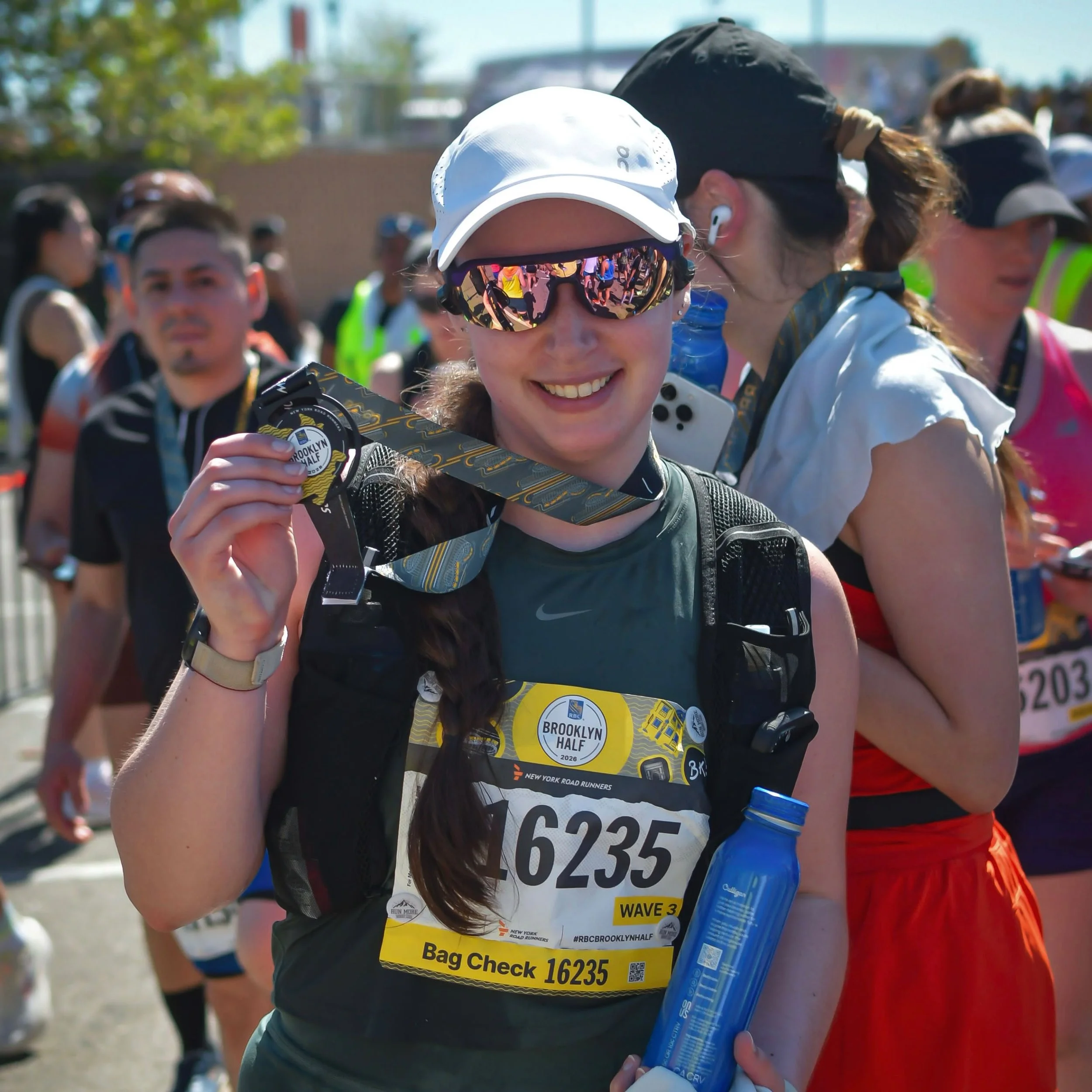 A woman running outdoors during a race, wearing a blue cap, black athletic tank top, black shorts, gray running shoes, and a black hydration vest, with a race bib attached to her top.