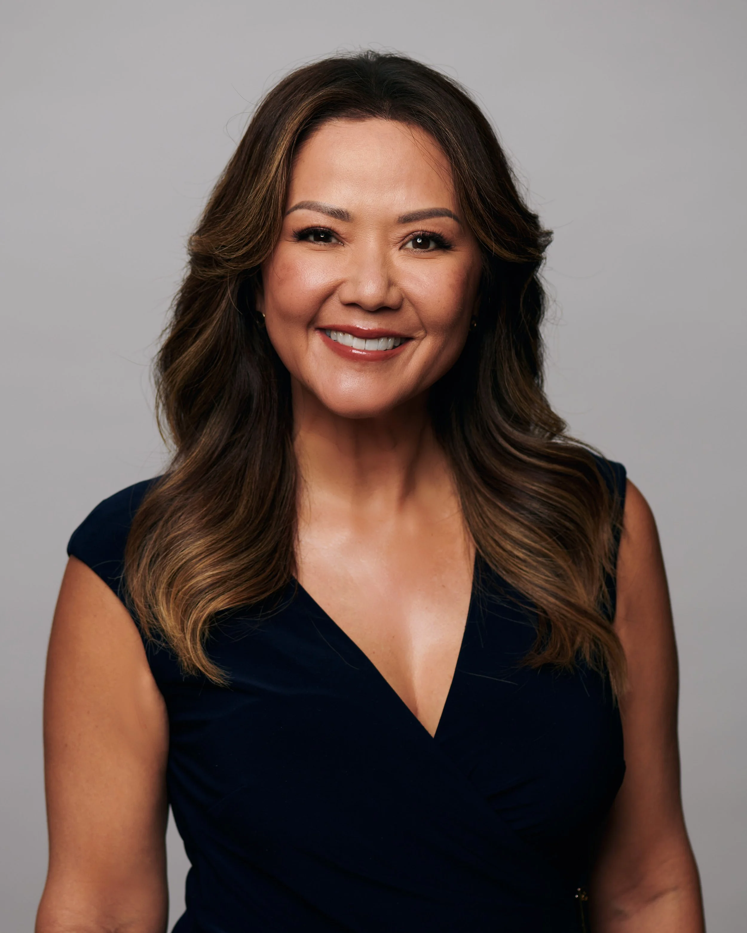 Headshot of a smiling woman with long, wavy brown hair, wearing a navy blue sleeveless top, against a plain gray background.