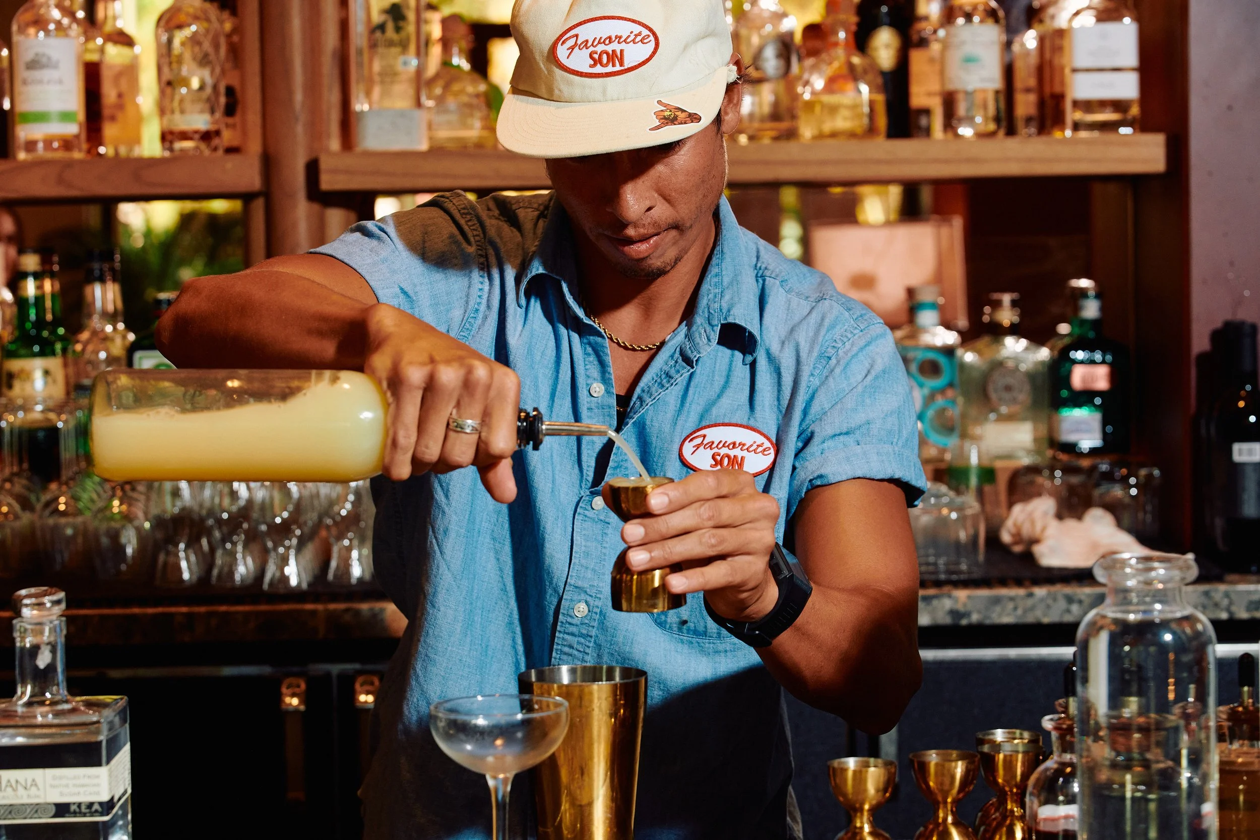 Bartender wearing a blue shirt and a beige cap pours a yellow cocktail into a glass at a bar with bottles and glasses in the background.