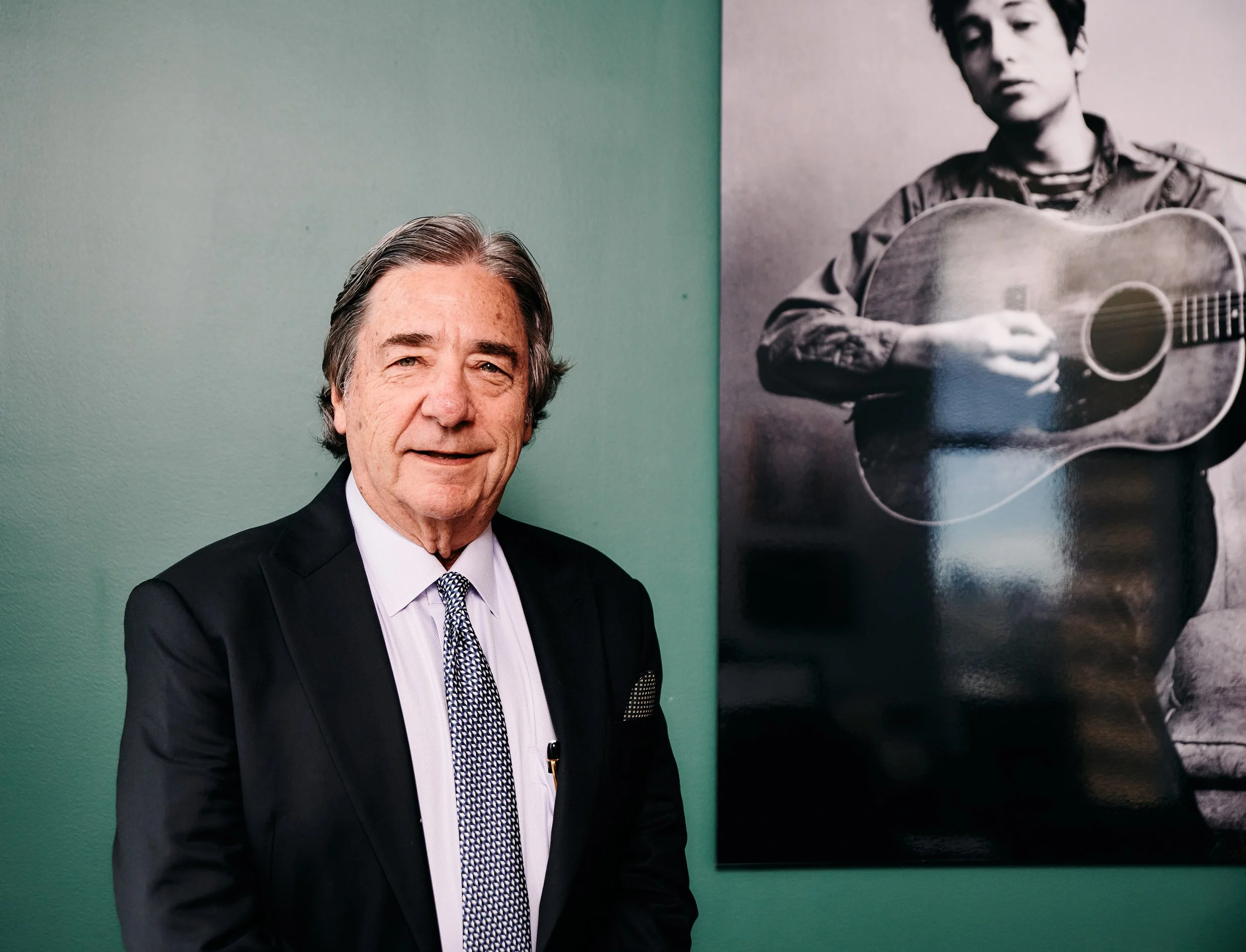 An elderly man in a black suit, white shirt, and patterned tie standing in front of a green wall with a black-and-white photograph of a young person playing a guitar.