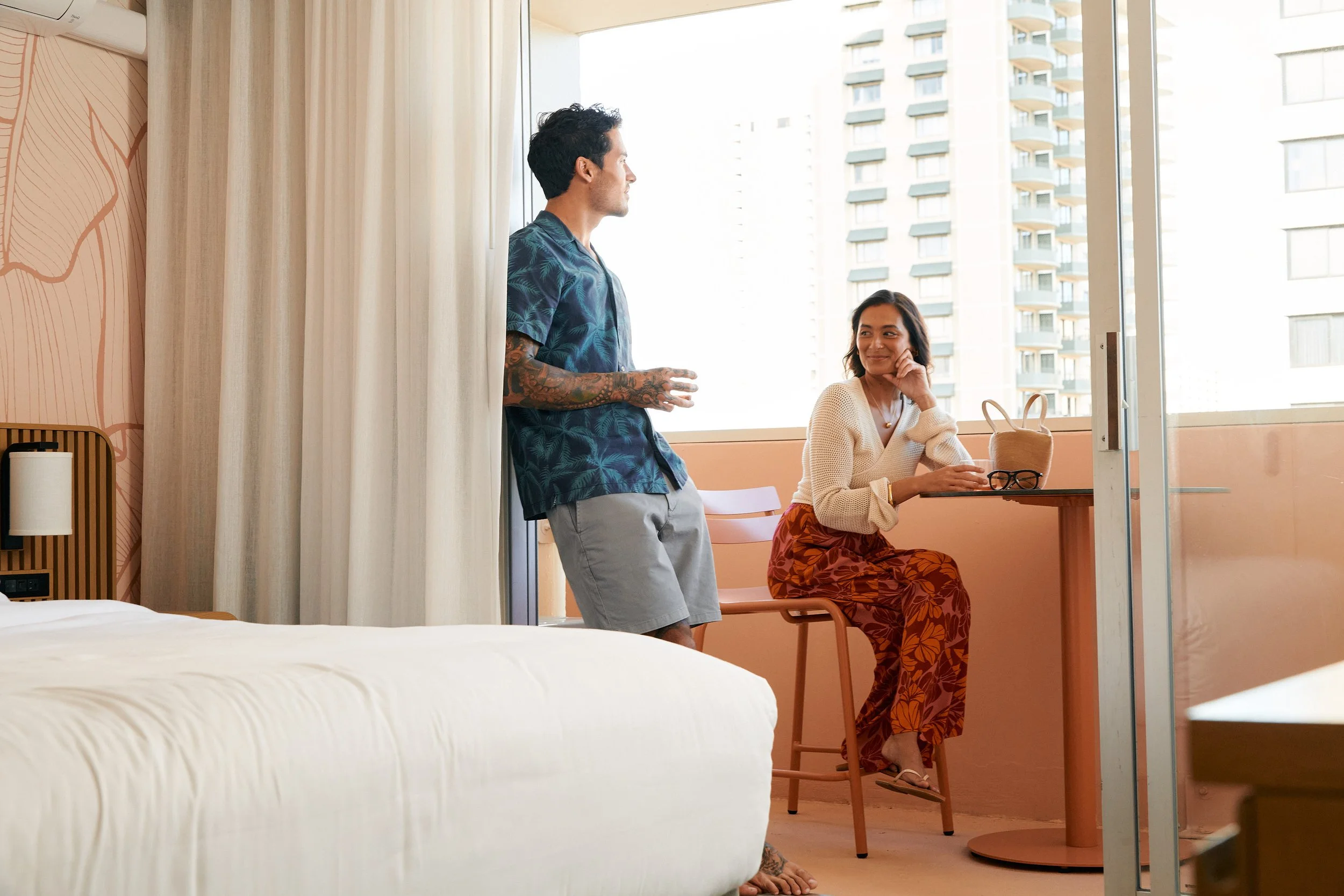 A man and woman in a bright hotel room. The woman is sitting at a small table near the window, smiling and looking at the man standing nearby. The man is leaning against the wall with arms crossed, talking to her. The room has natural light, a bed in