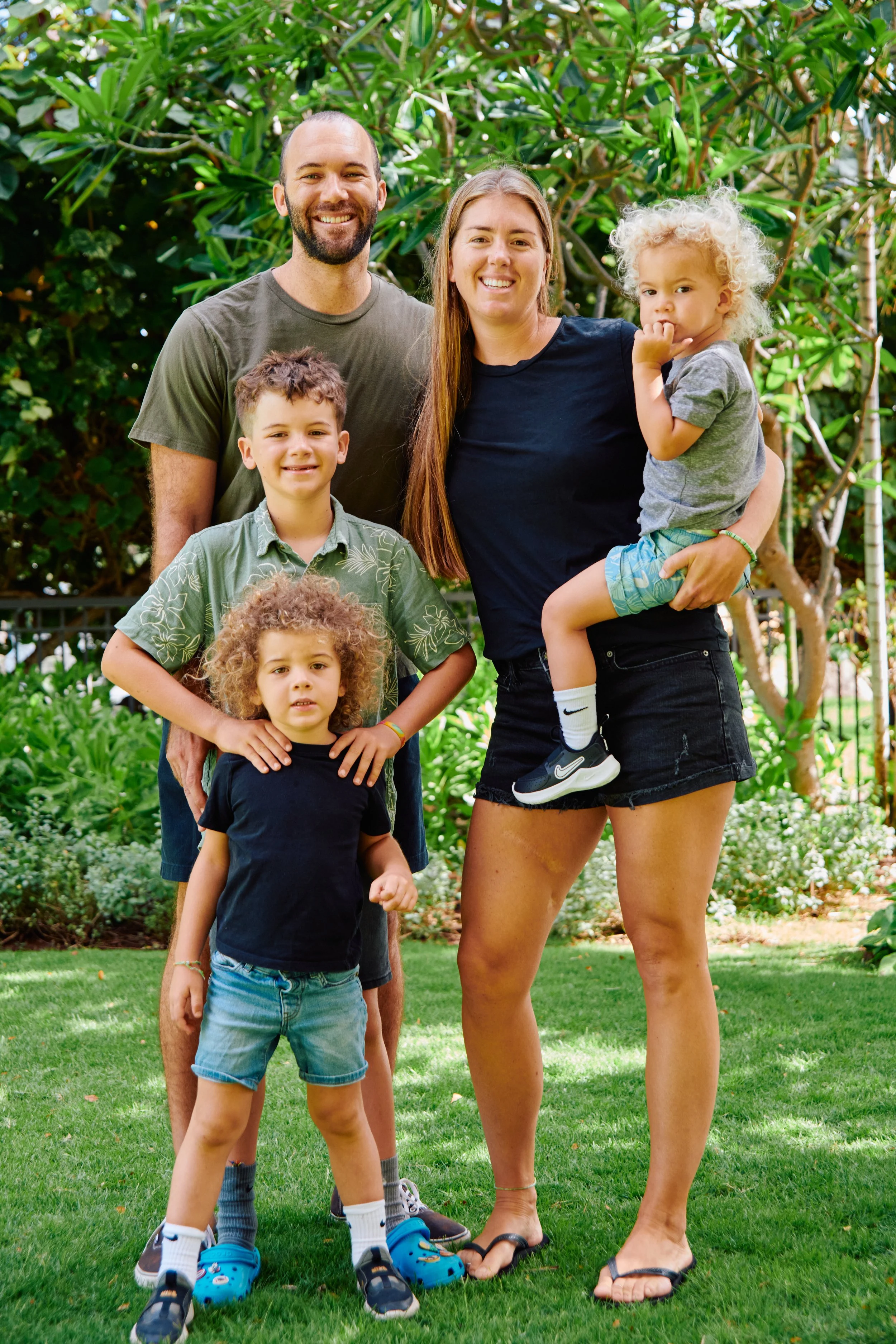 A family of five standing together outdoors in a garden with lush green foliage. The adults, a man and a woman, are smiling while holding their children. One child is standing in front of the father, and a younger girl is being held by the mother.