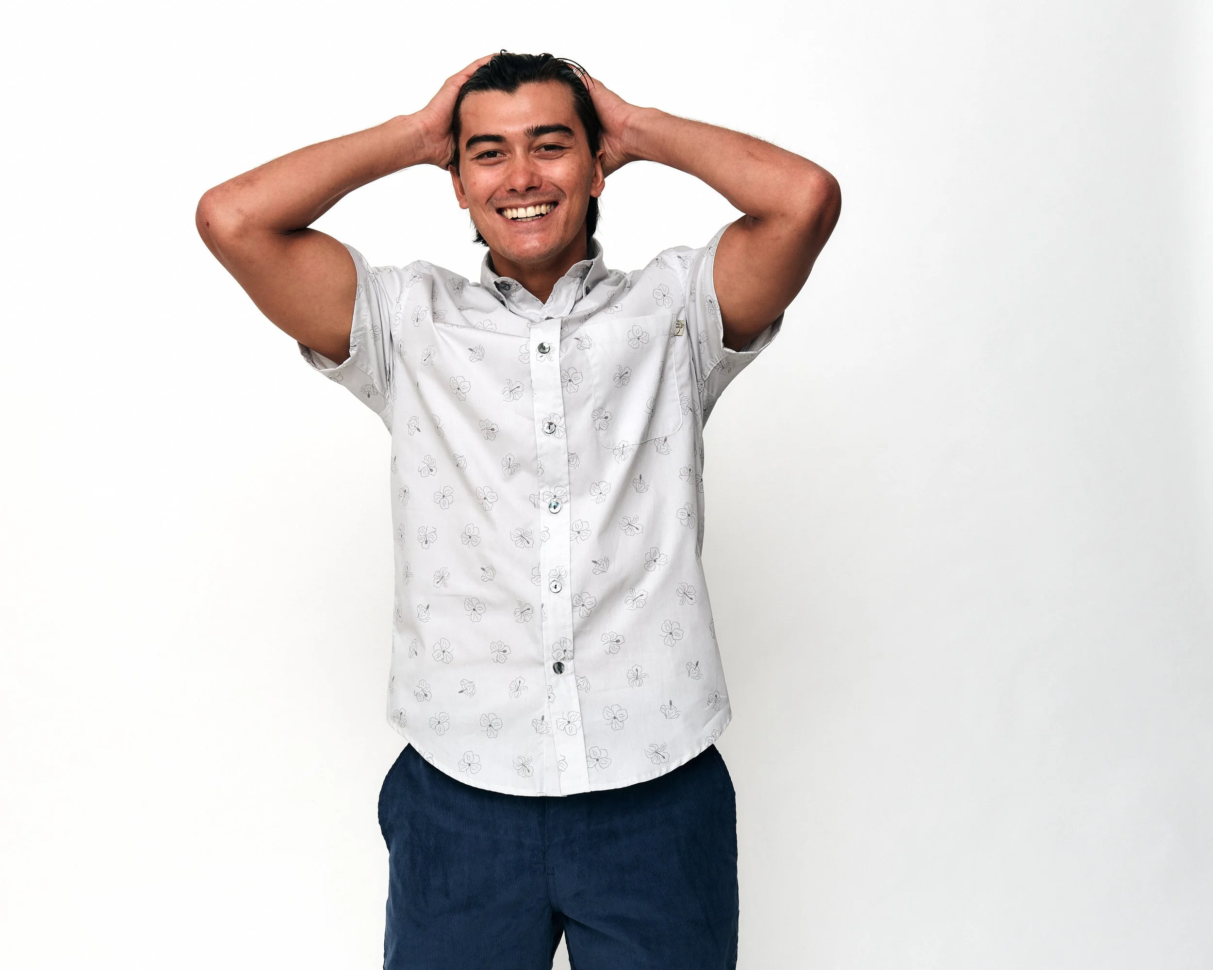 Young man smiling with hands on head, wearing a white short-sleeve shirt with a butterfly pattern and dark blue pants, standing against a plain white background.