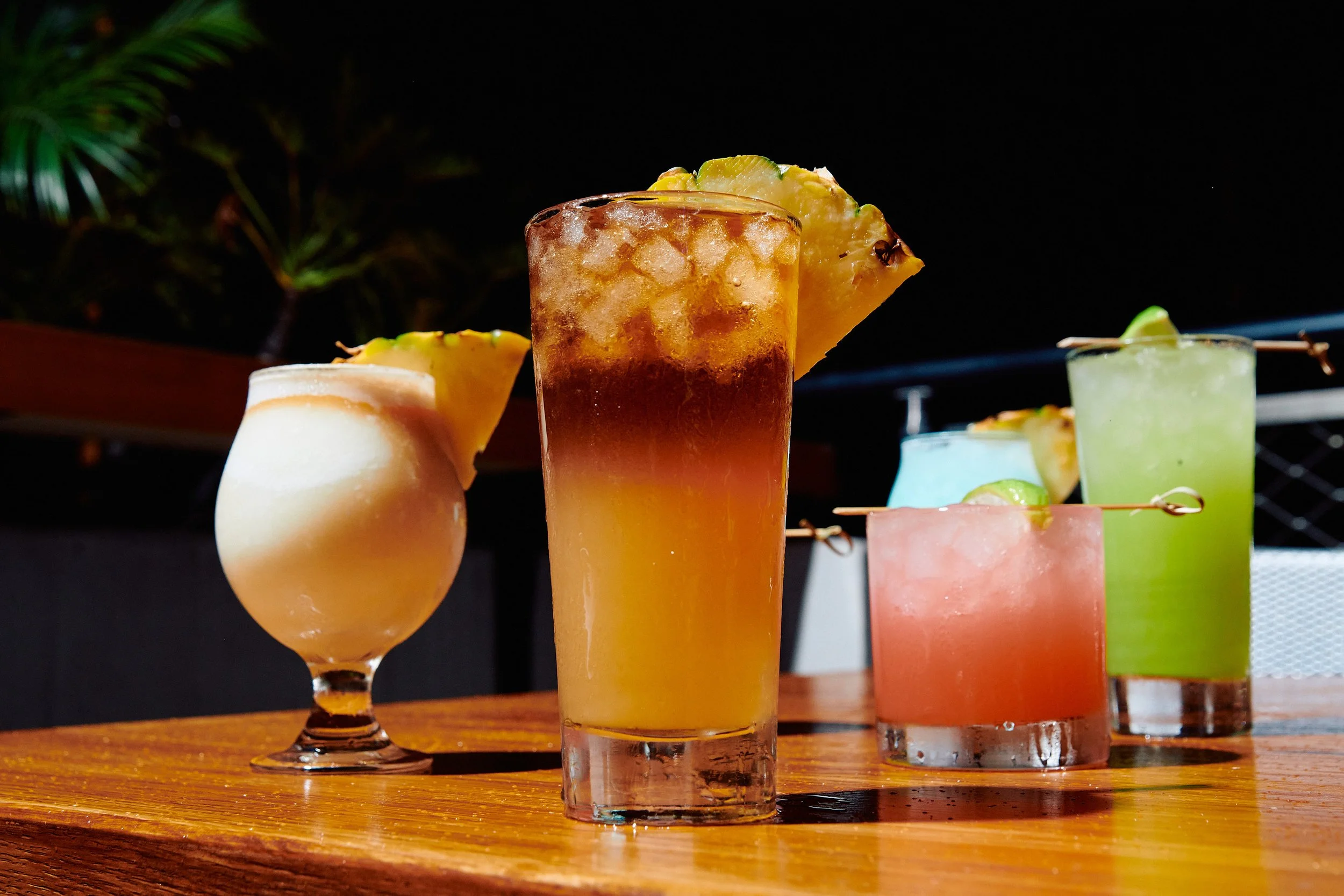 Assorted colorful tropical cocktails with pineapple garnish on a wooden surface at night.