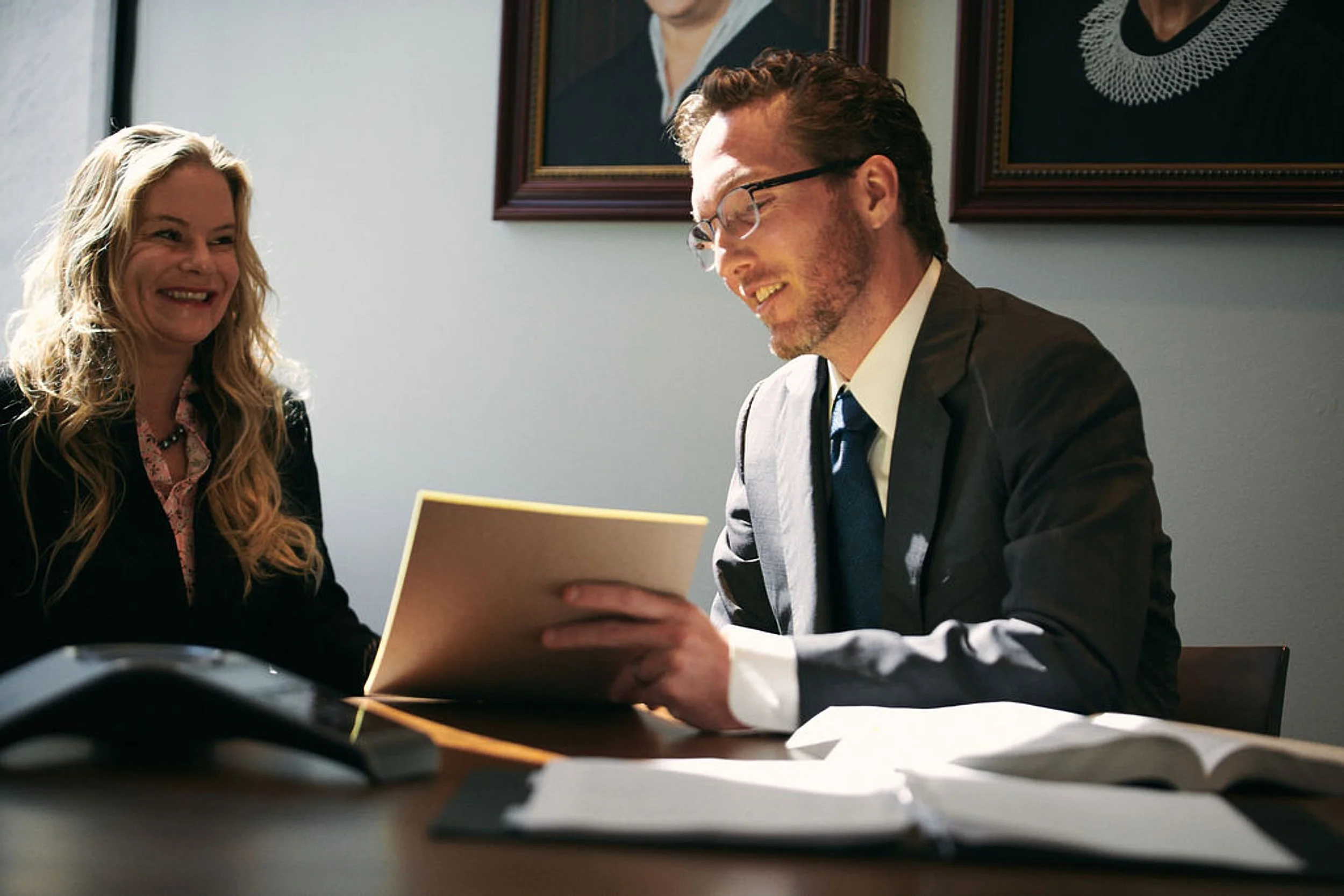 A man and woman sitting at a desk, engaging in a discussion with documents and a phone on the table, in a professional office setting.