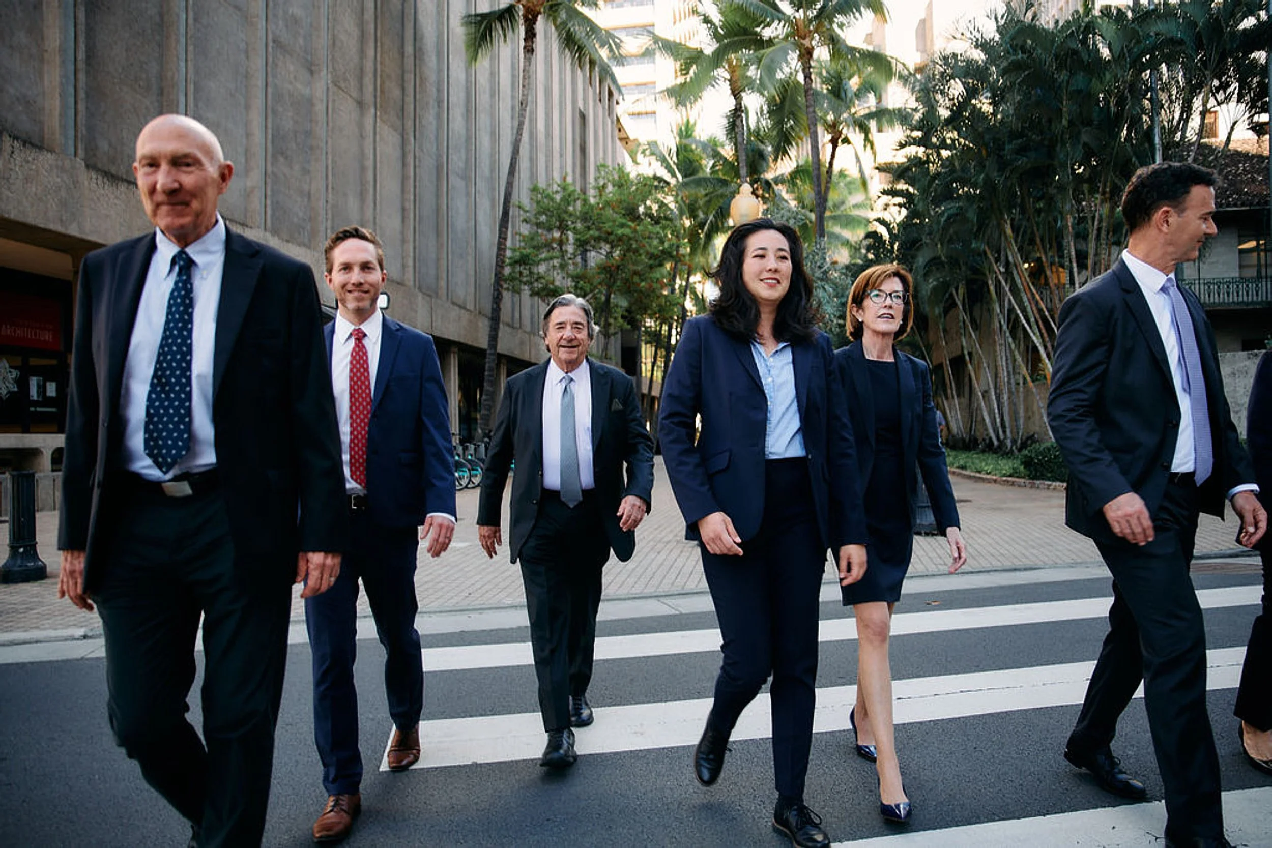 Group of professionally dressed people walking across a crosswalk in an urban area with trees and buildings in the background.