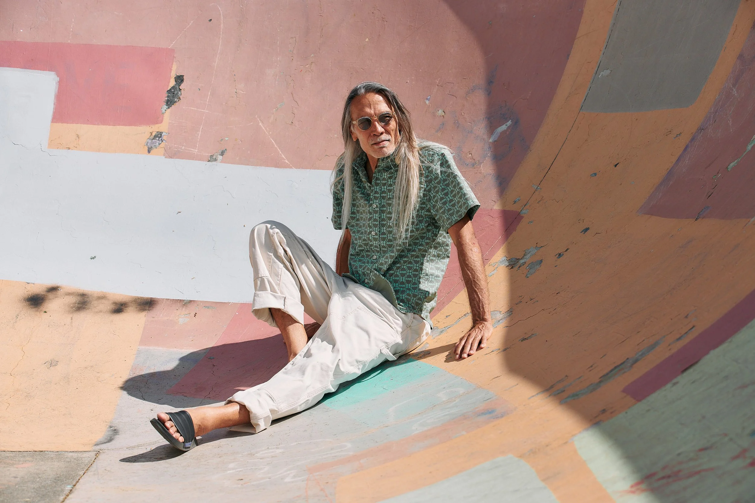Man sitting on the ground in a colorful skate park or concrete structure, wearing sunglasses, a patterned green shirt, and beige pants.