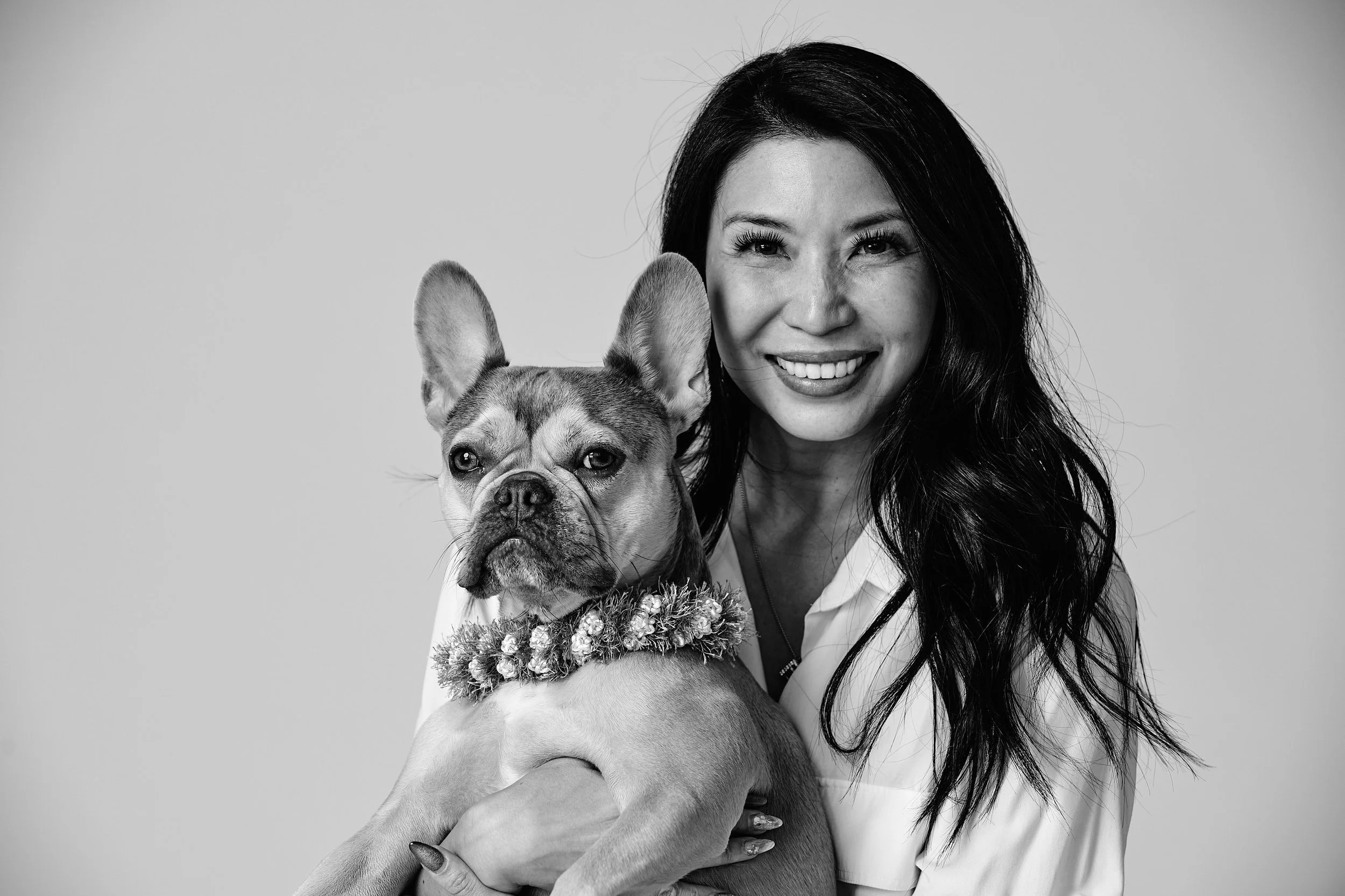 Black-and-white photo of a woman with long, dark hair smiling and holding a French Bulldog wearing a floral collar.