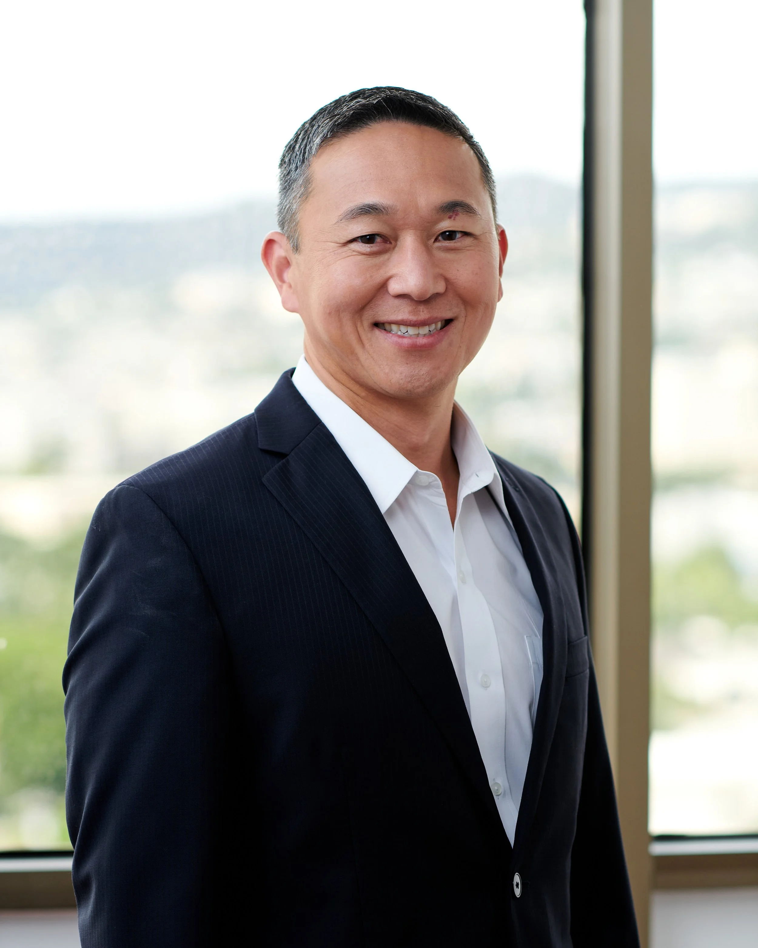 Portrait of a smiling man in a dark suit and white shirt standing in front of a window with a cityscape background.