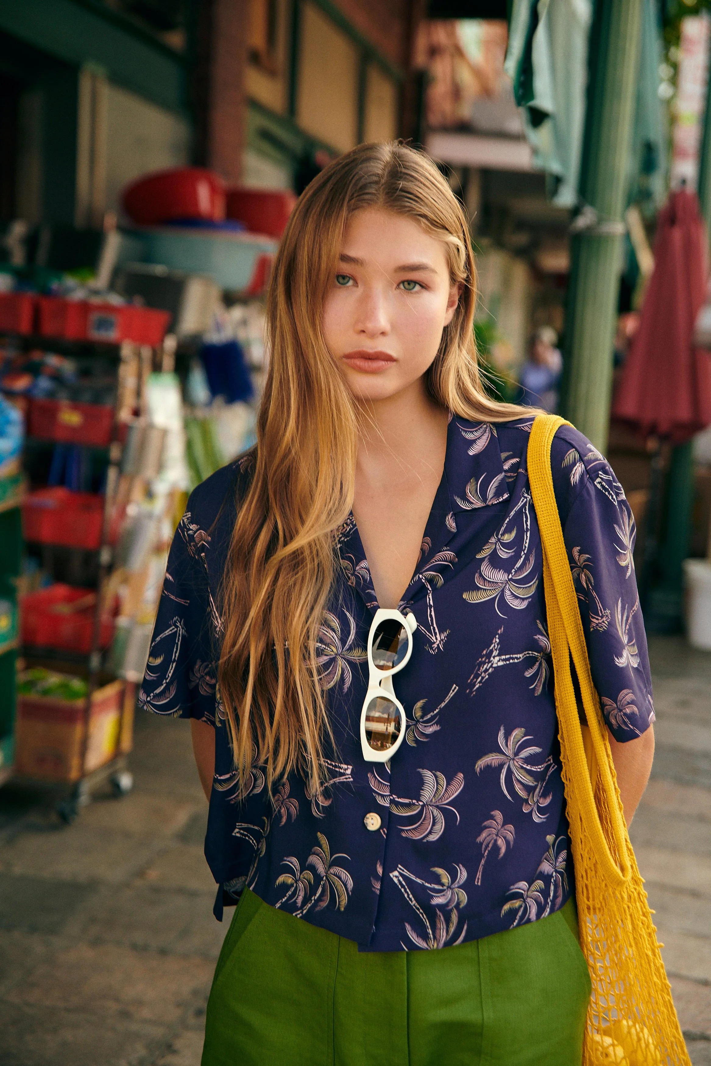 A young woman with long, wavy blonde hair standing outdoors in a market or street fair setting, wearing a navy blue floral patterned shirt, green pants, and carrying a yellow netted bag over her shoulder, with a pair of white sunglasses hanging from 