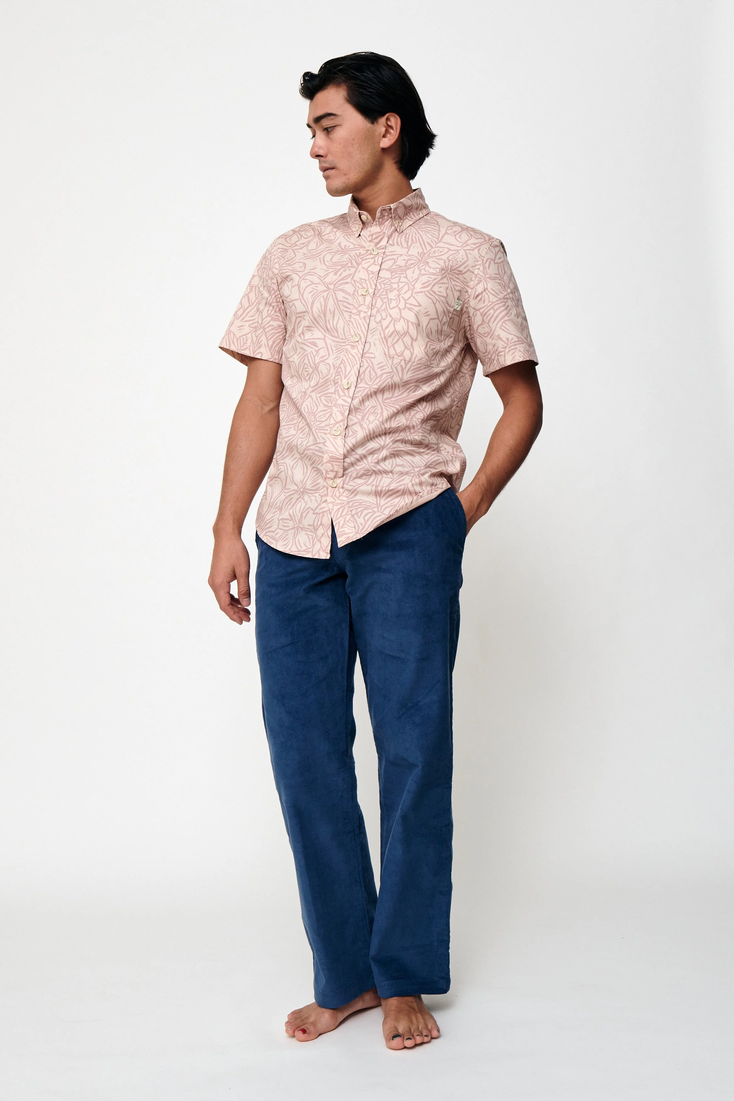 A man standing barefoot against a plain white background, wearing a short-sleeved, button-up shirt with a floral pattern and dark blue pants.