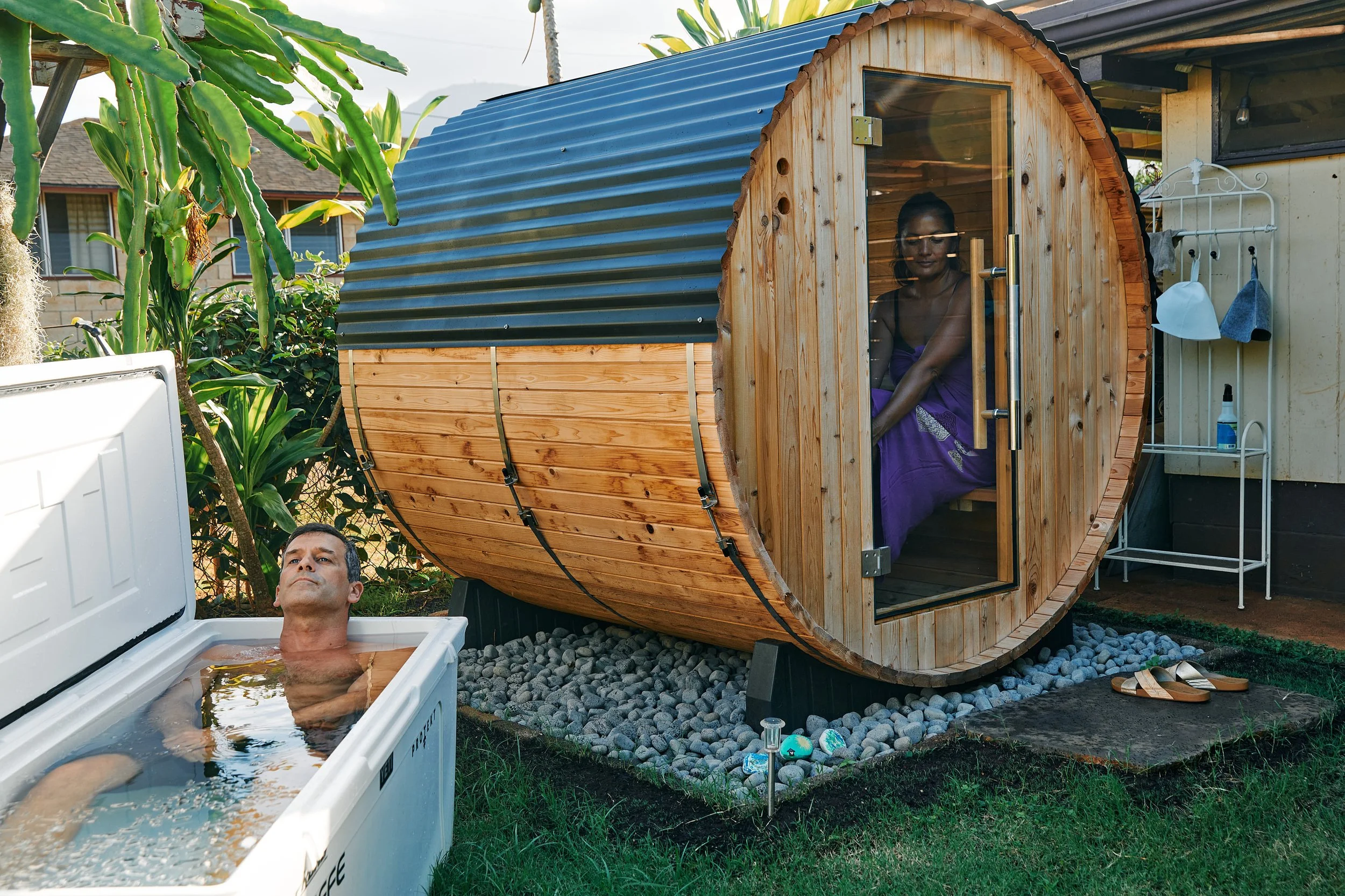A man relaxing in an outdoor hot tub and a woman sitting inside a barrel sauna in a backyard surrounded by plants.