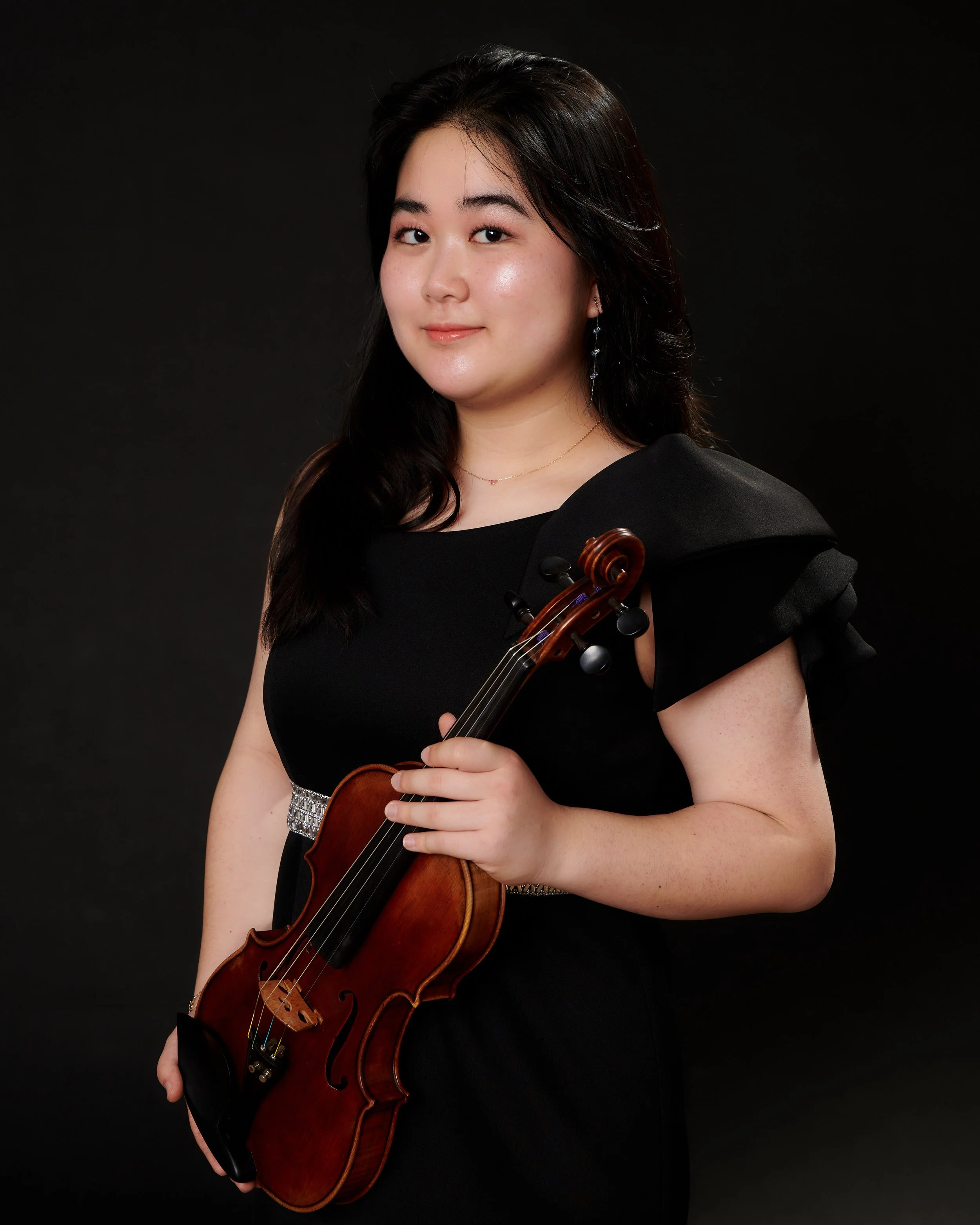 A woman in a black dress holding a violin, standing against a black background.
