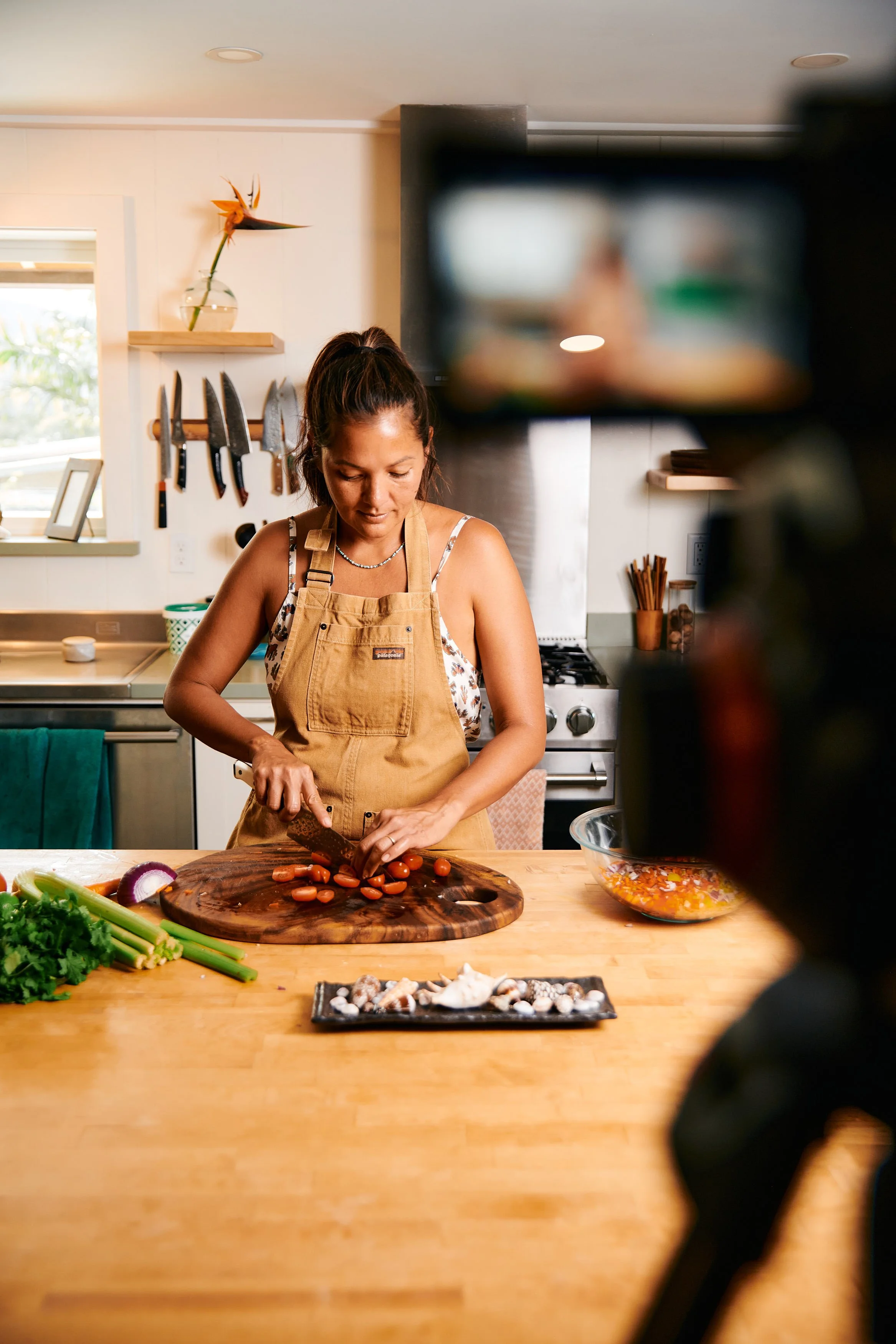 A woman chopping carrots on a wooden cutting board in a kitchen, with vegetables and cooking ingredients around her, as a camera captures her preparing a meal.