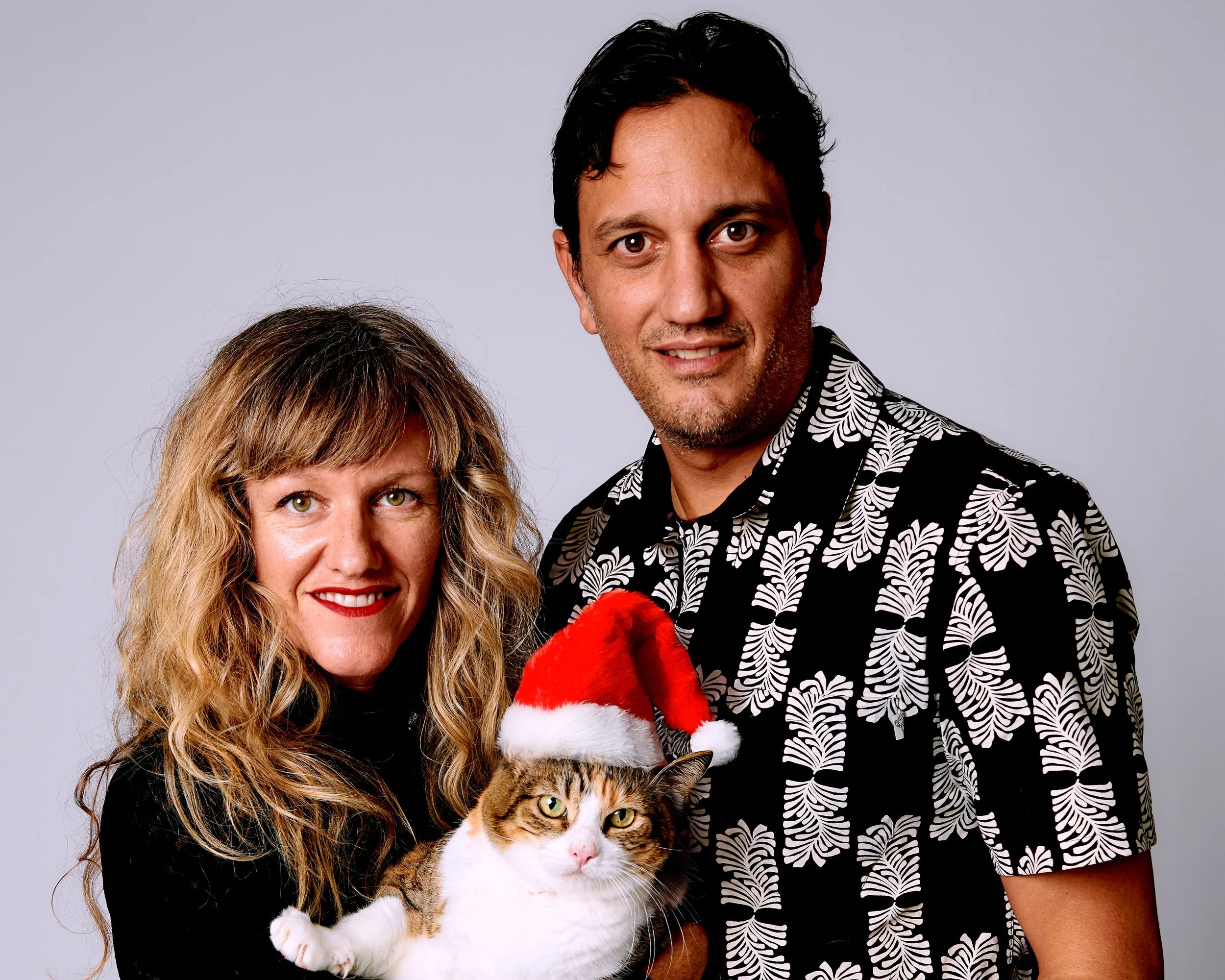 A smiling woman with wavy hair, a smiling man with dark hair, and a cat wearing a Santa hat, against a plain gray background.