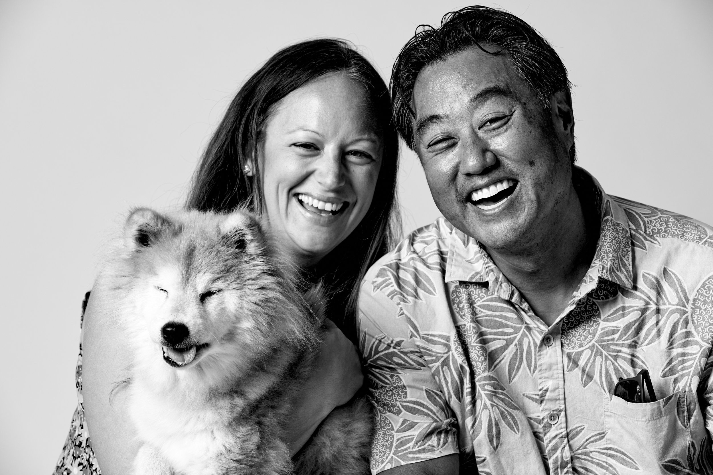 A smiling woman and a smiling man with a fluffy dog, in black and white.