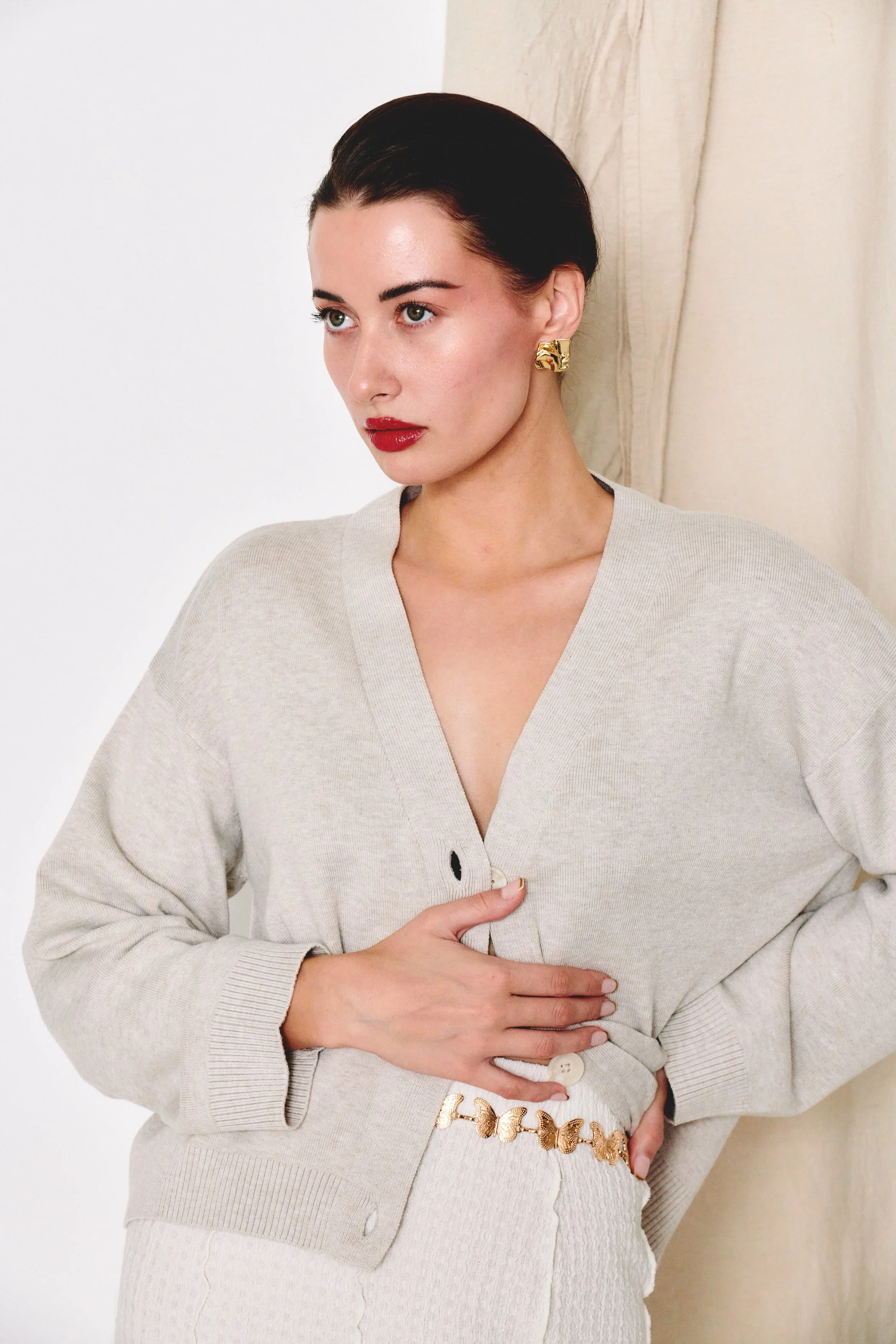 A woman with dark hair and red lipstick is wearing a beige cardigan and jewelry, standing against a neutral background.