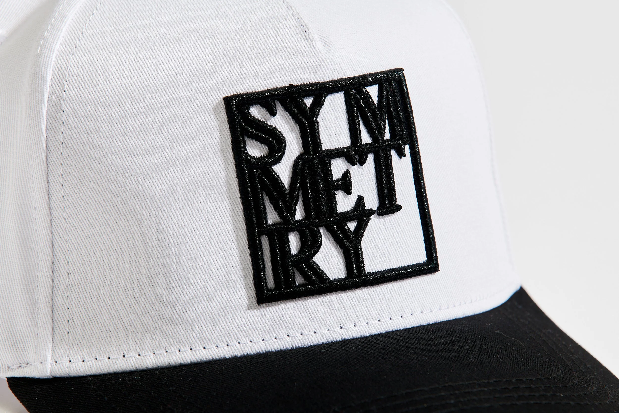 A baseball cap with a white crown, black brim, and a black embroidered patch on the front that reads 'SYLM REY' in stylized text.