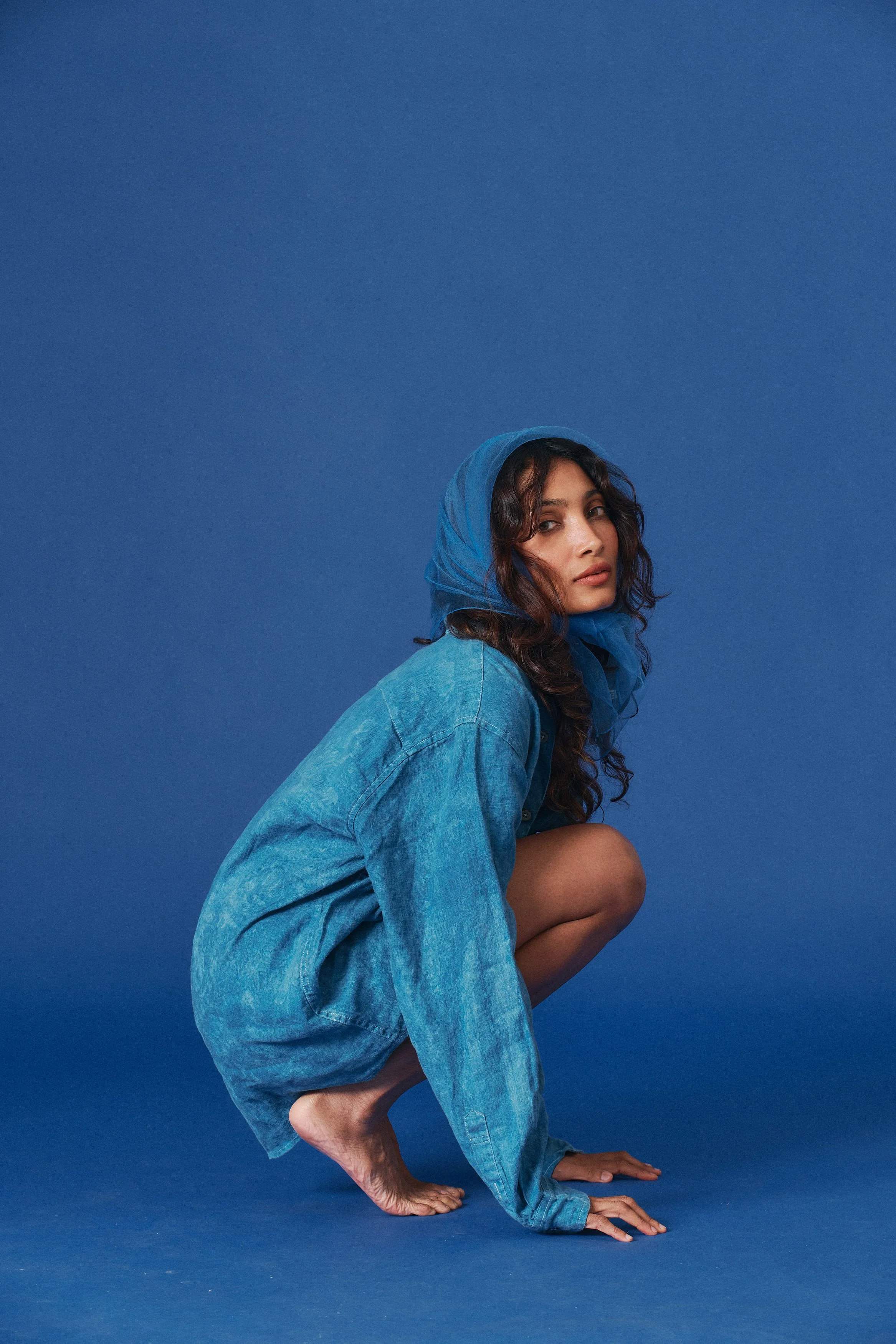 A woman crouching on her hands and feet against a blue background, wearing a loose denim jacket and a matching blue headscarf.