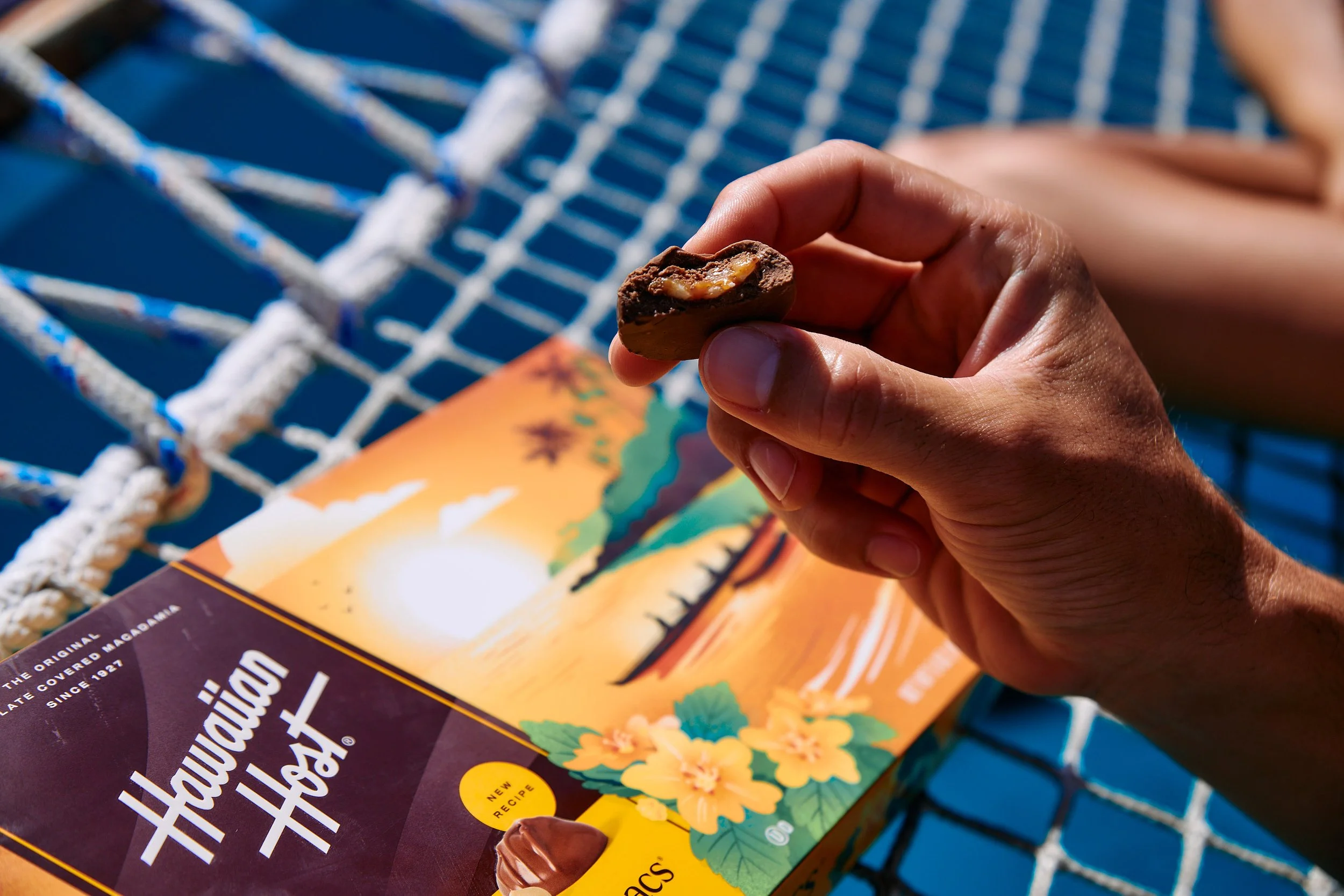 A person holding a chocolate confection with caramel inside, sitting on a blue outdoor surface with a straw woven pattern and a colorful magazine showing a sunset scene and hibiscus flowers.