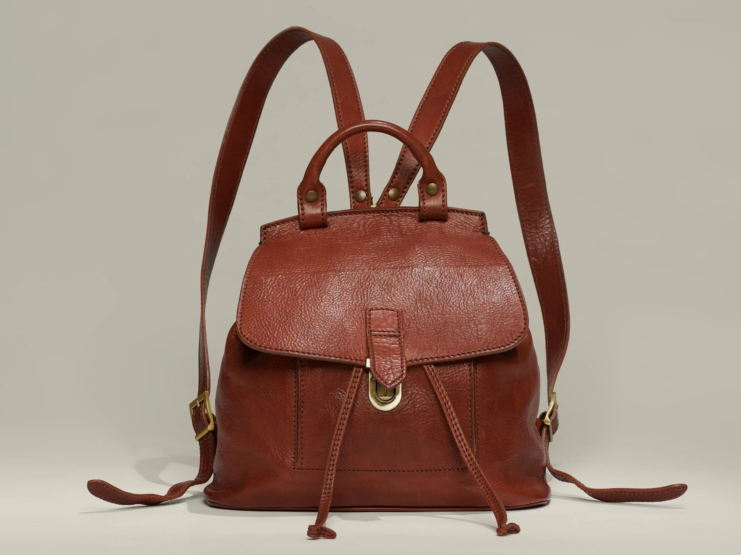 A brown leather handbag with shoulder straps and a top handle, featuring a small front flap with buckle detail, against a neutral background.