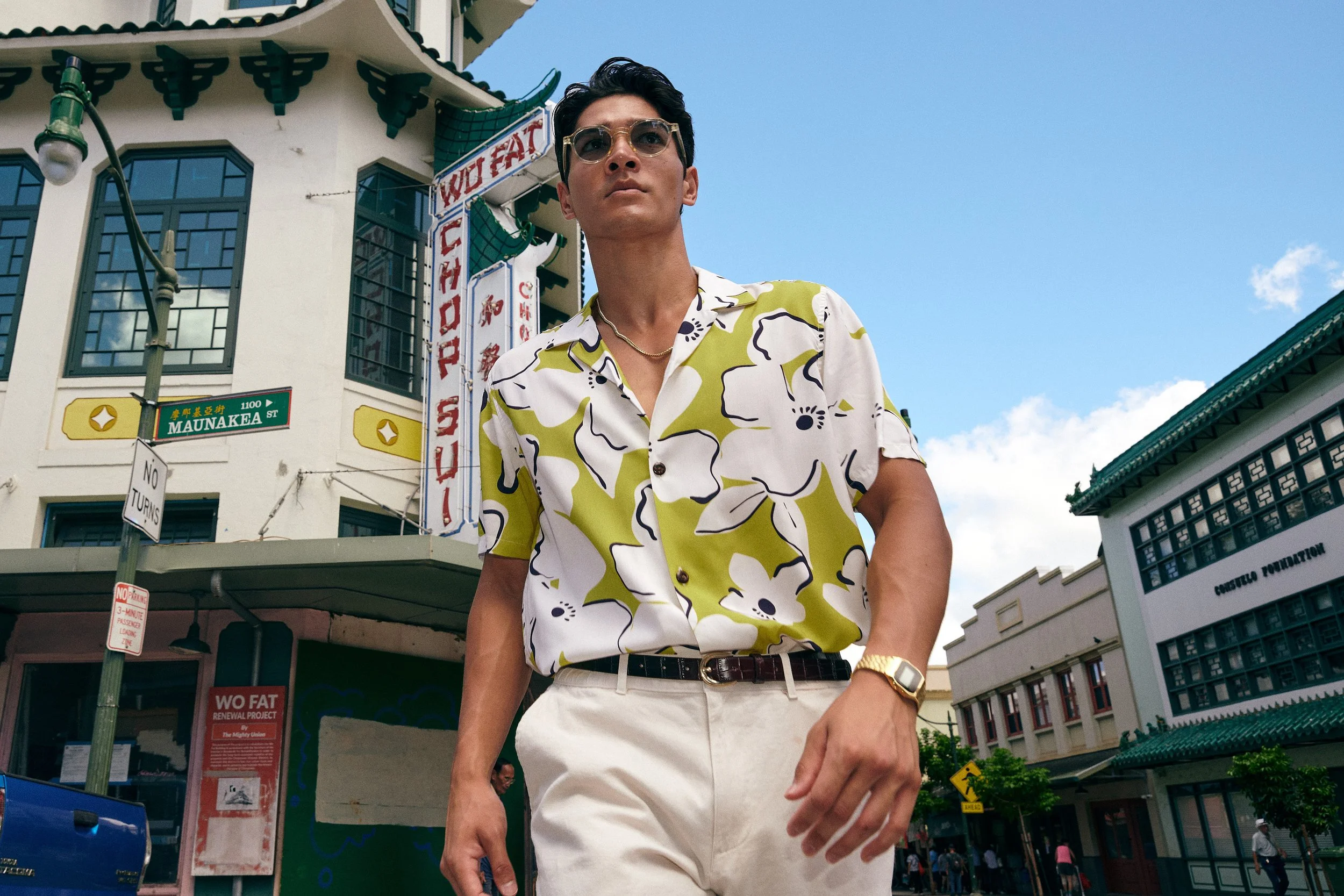 A man wearing sunglasses and a floral shirt walking on a city street with buildings and signs behind him, including a street sign for Maunakea street and a building with a sign that reads 'East Chinatown'.